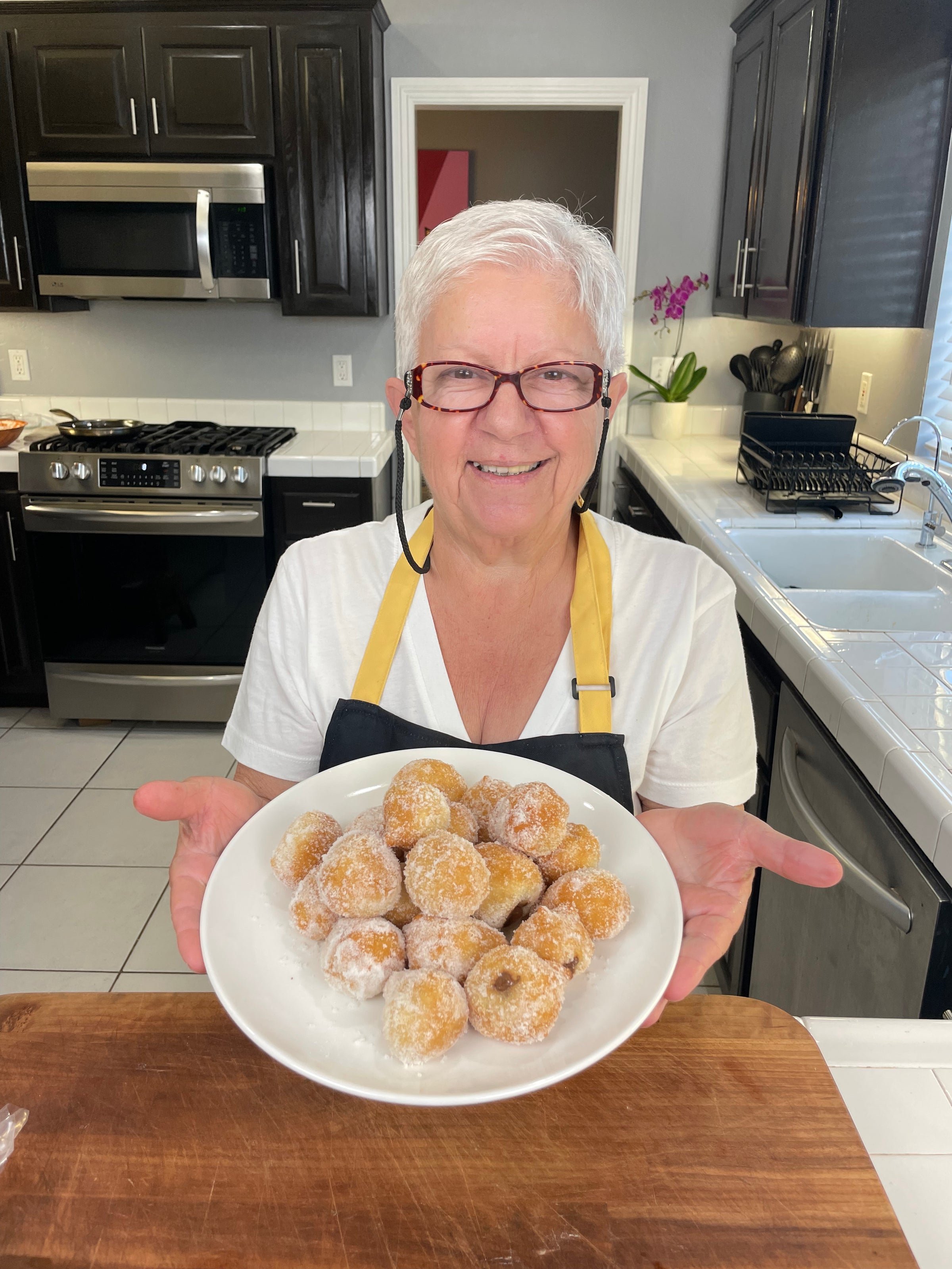 Smiling older woman with short white hair and glasses, wearing a yellow apron, holds a plate of round, sugar-coated pastries in a modern kitchen with dark cabinets and stainless steel appliances.