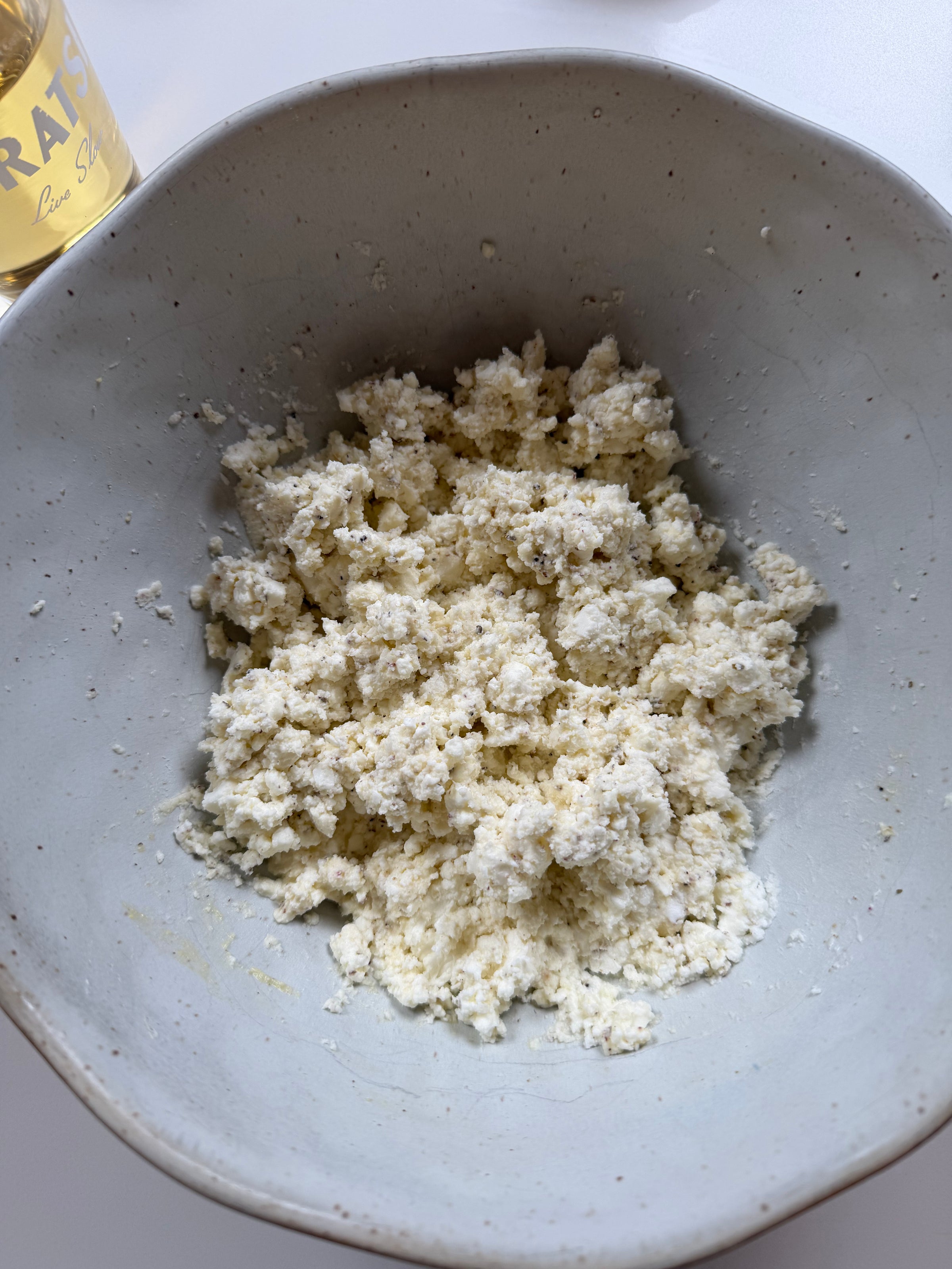 A close-up of a grey ceramic bowl containing crumbled ricotta cheese. In the top left corner, part of a glass bottle labeled GRAYS is visible. The bowl sits on a white surface.