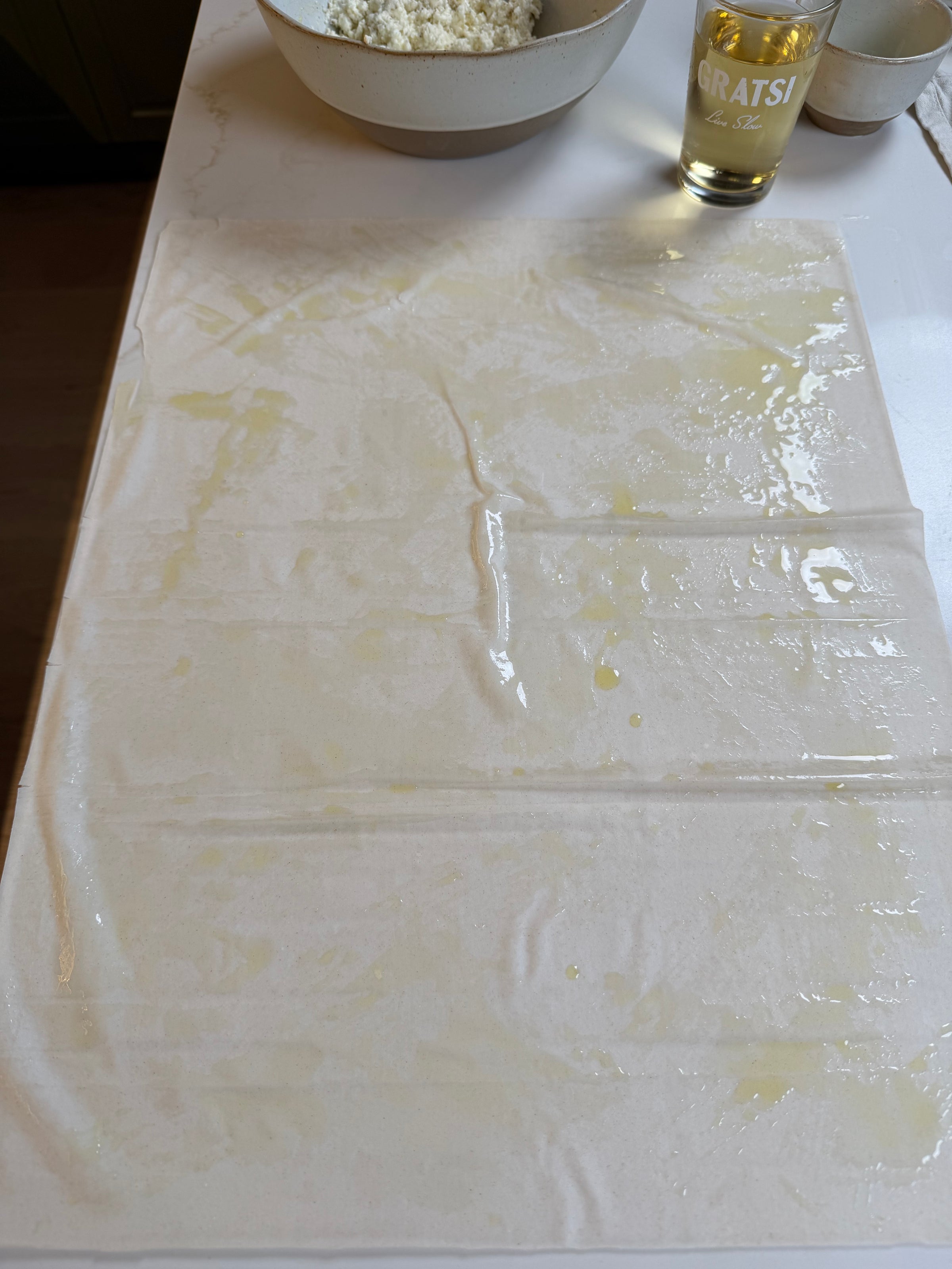 A sheet of filo dough is spread out on a white surface and brushed with oil. Nearby are a bowl filled with crumbled cheese and a glass labeled GRATSI partially filled with a light-colored liquid.