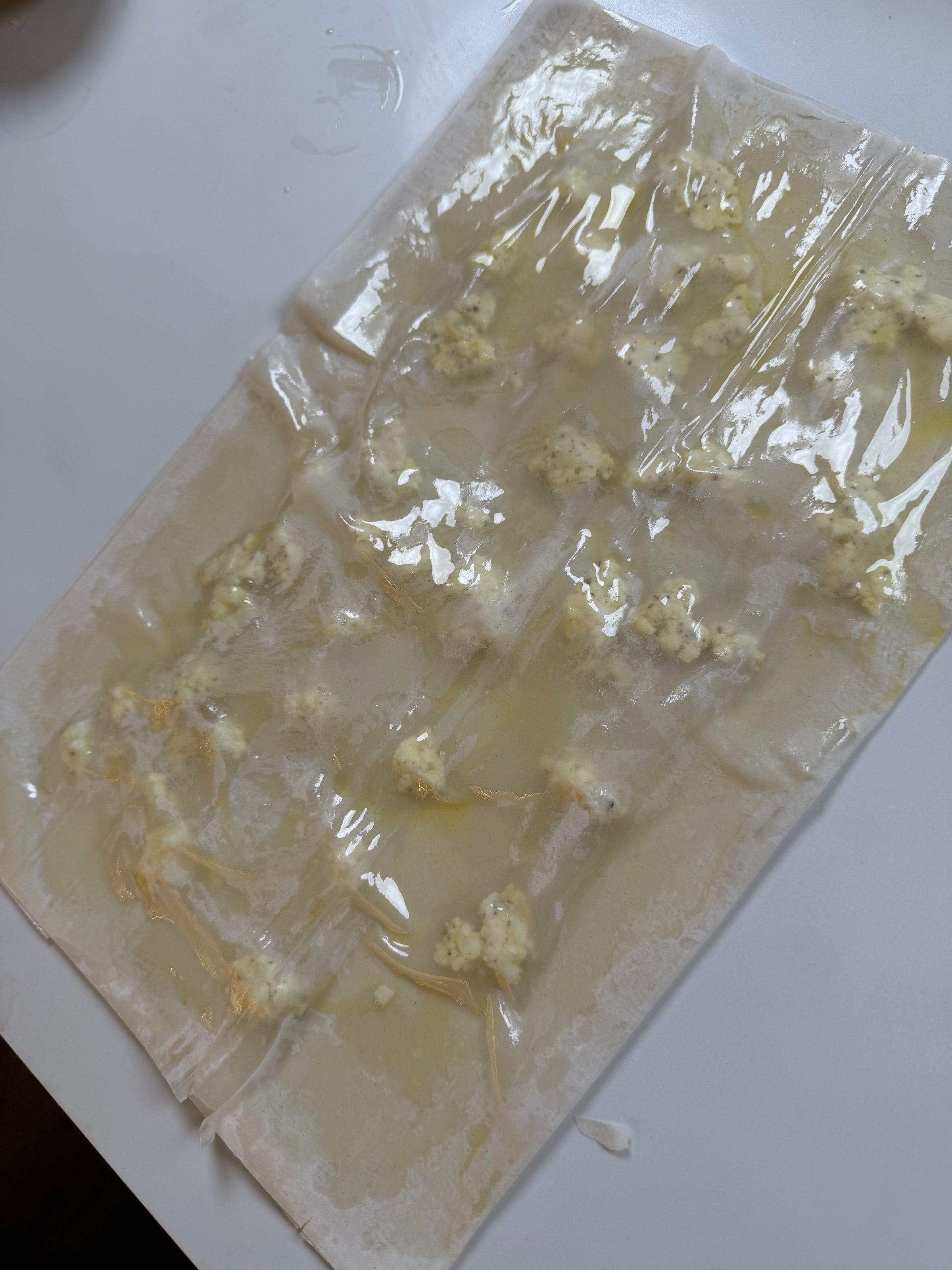 A sheet of pasta dough on a white surface is dotted with small mounds of creamy ricotta cheese filling, with some egg wash or water brushed around the dollops, likely in preparation for homemade ravioli.