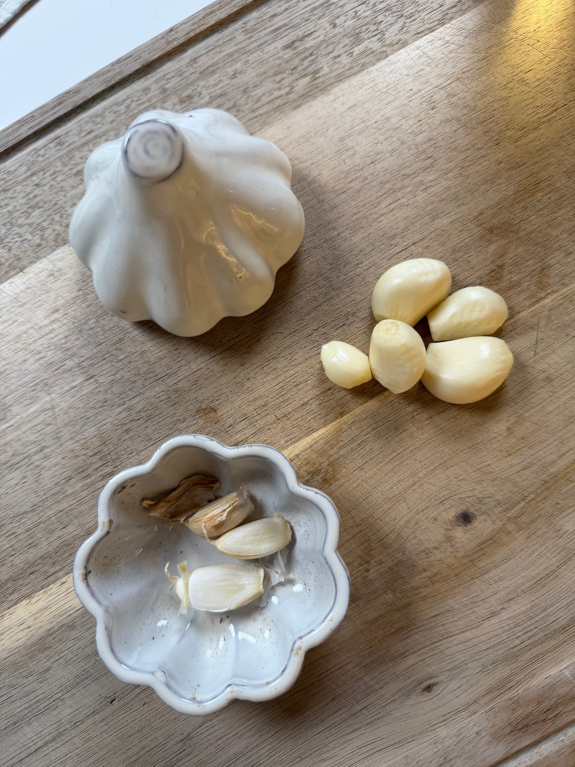 A whole garlic bulb, several peeled garlic cloves, and a small white dish with garlic skins sit on a wooden surface.