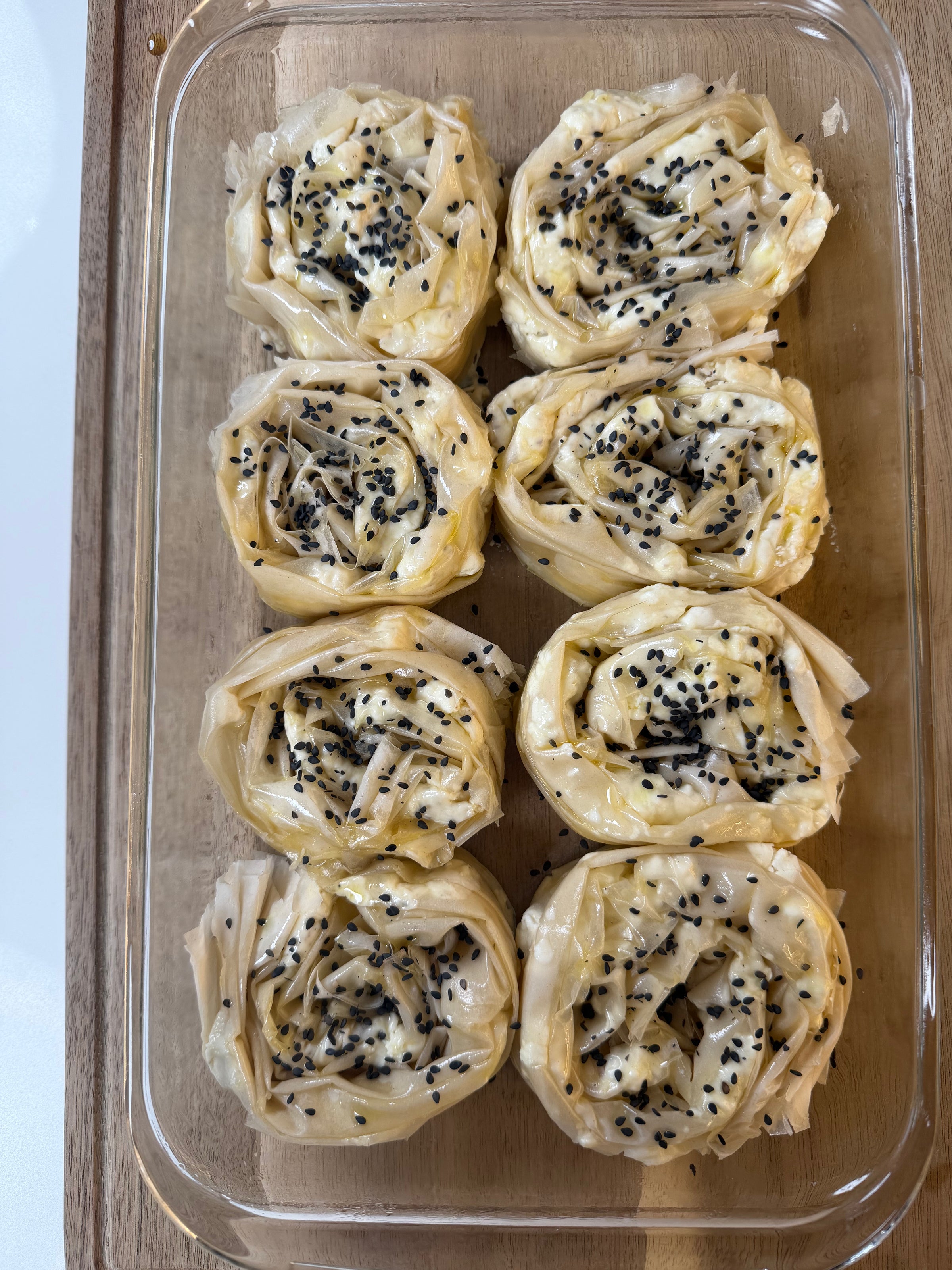 A glass baking dish holds seven rolled pastries made with thin dough, filled with a white cheese and sprinkled with black sesame seeds, arranged neatly in two rows on a wooden surface.