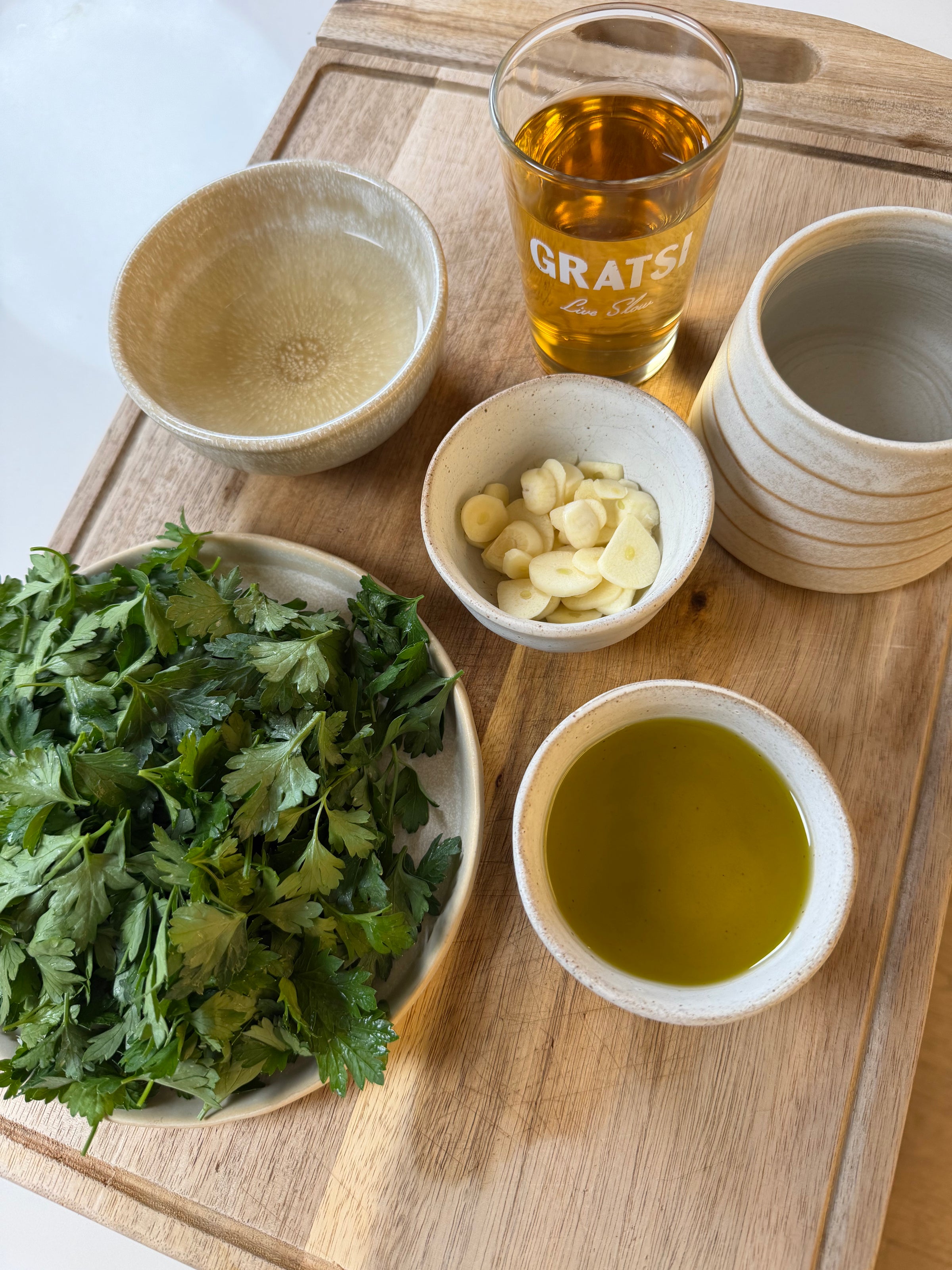 Bowls with chopped cilantro, sliced garlic, olive oil, and an empty bowl are on a wooden board. A glass labeled GRATSI contains a golden liquid, possibly vinegar or wine, next to the other ingredients.