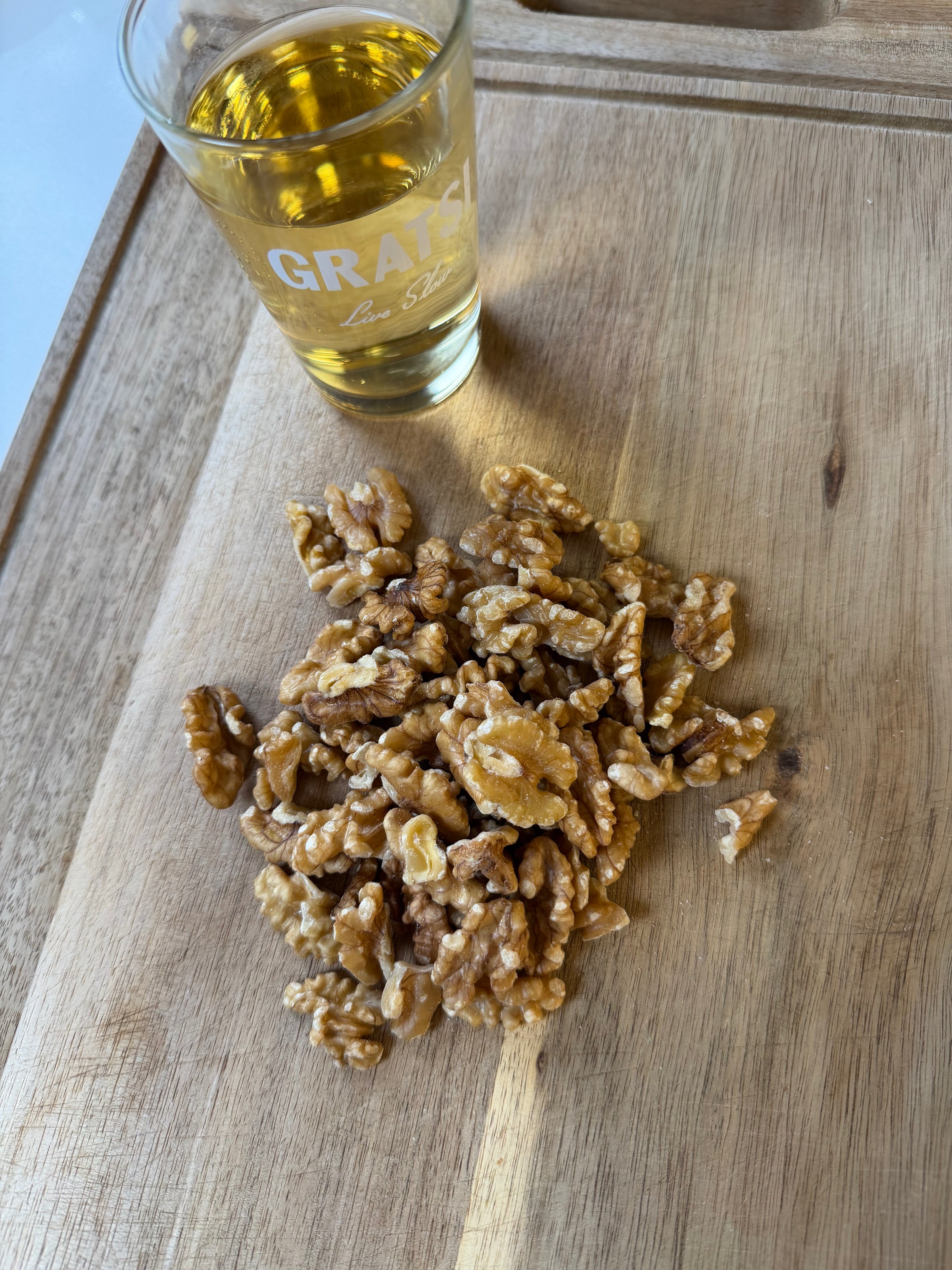 A glass of light yellow liquid stands on a wooden surface next to a pile of shelled walnut halves.