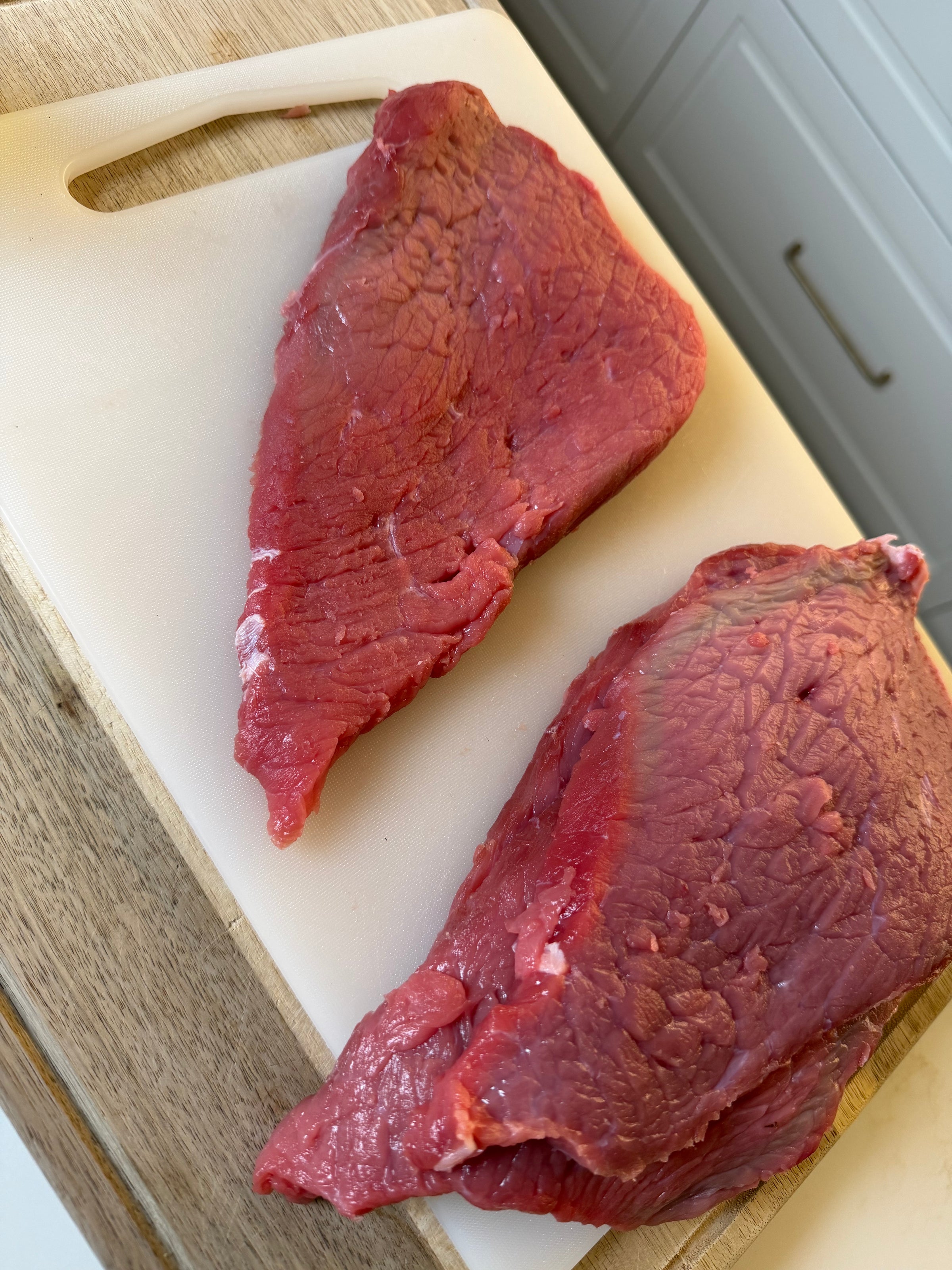 Two raw beef steaks sit on a white cutting board, which is placed on a wooden surface. Kitchen drawers and cabinets are visible in the background.