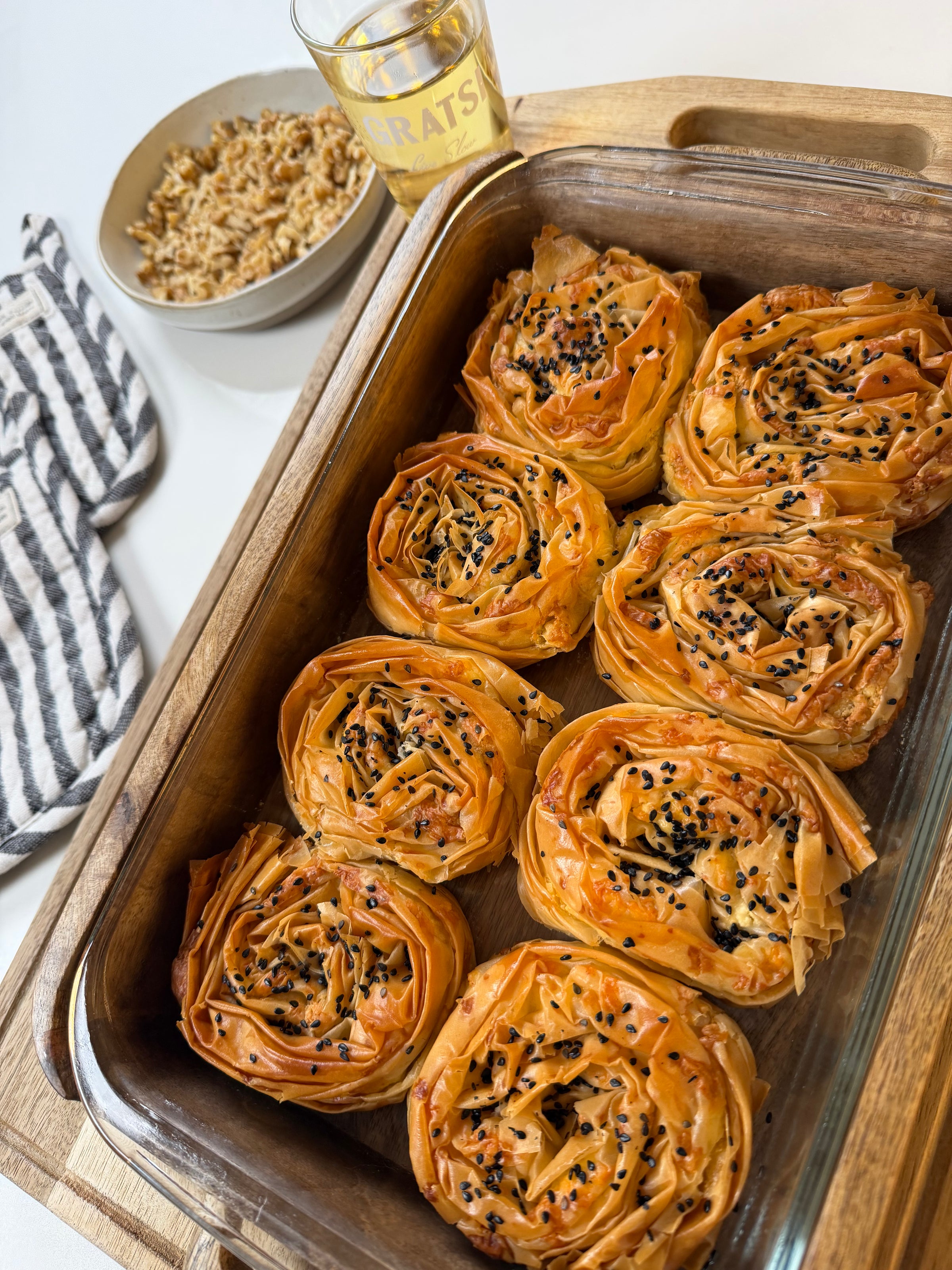 A baking dish with seven golden, spiral-shaped pastries topped with black sesame seeds, next to a striped towel, a bowl of nuts, and a glass with “CONGRATS” written on it.