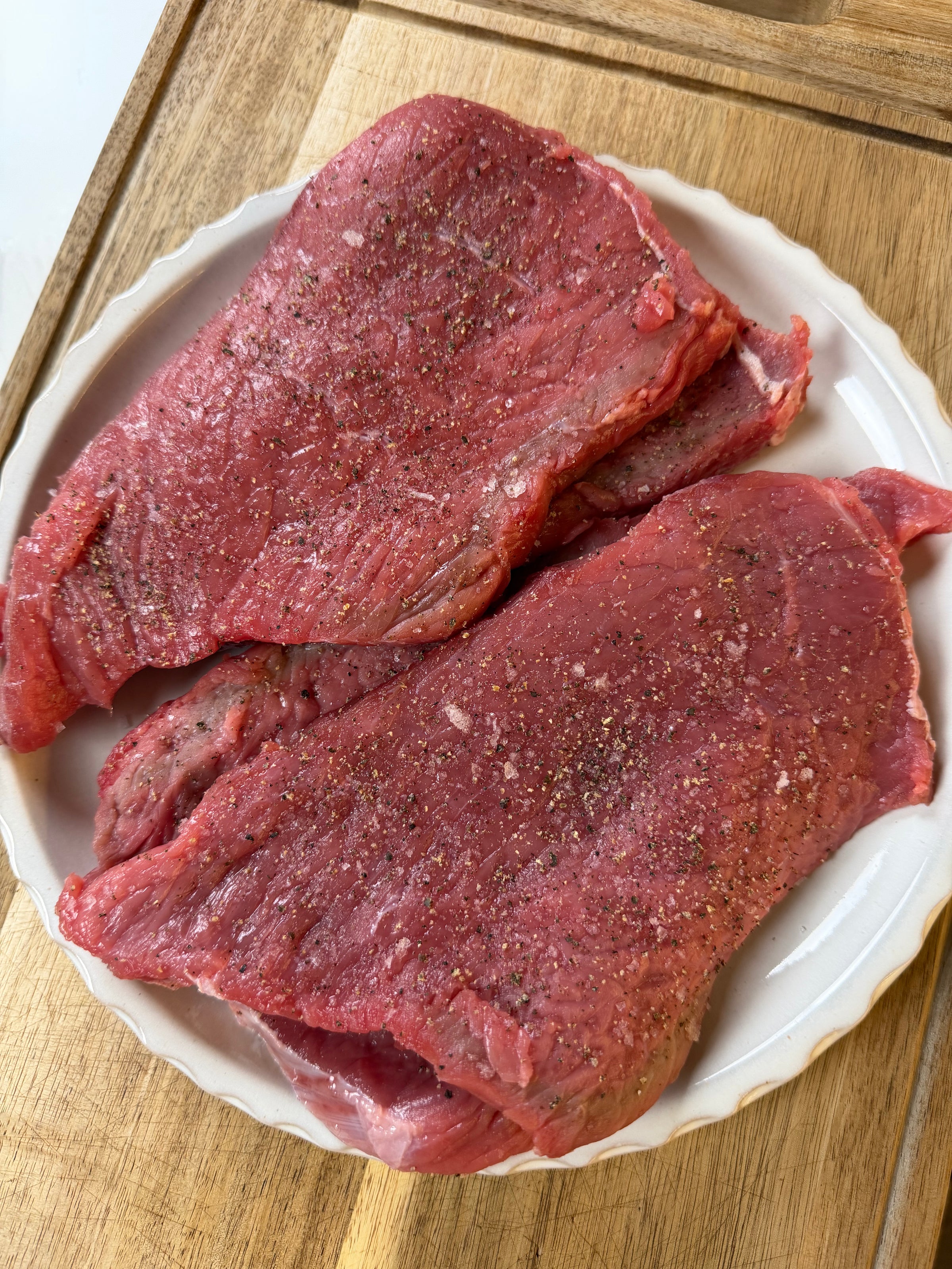 Two raw beef steaks seasoned with salt and pepper sit on a white plate placed on a wooden cutting board.
