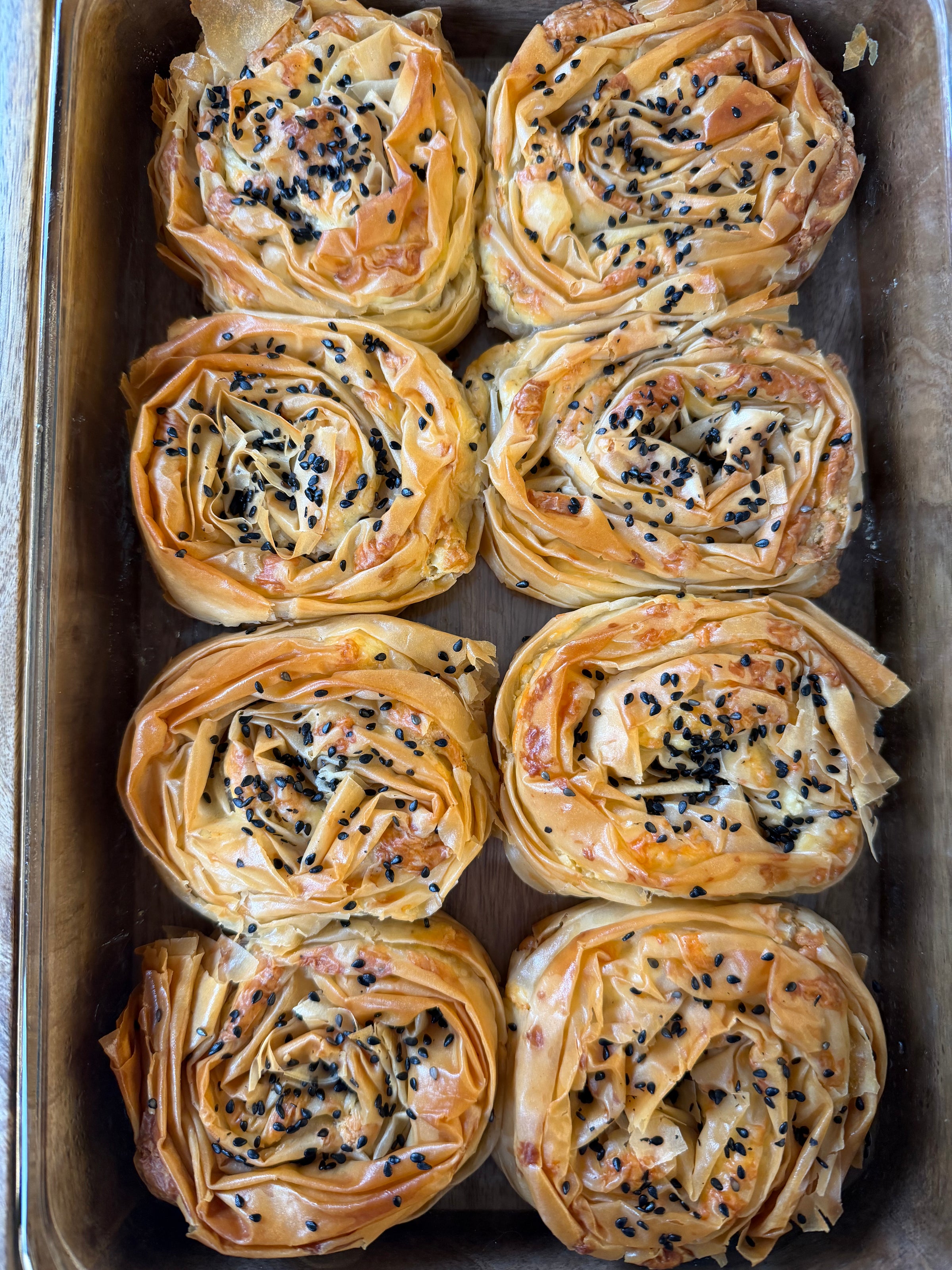 Eight golden, spiral-shaped pastries with crispy phyllo layers and black sesame seeds on top are arranged neatly in a baking tray.