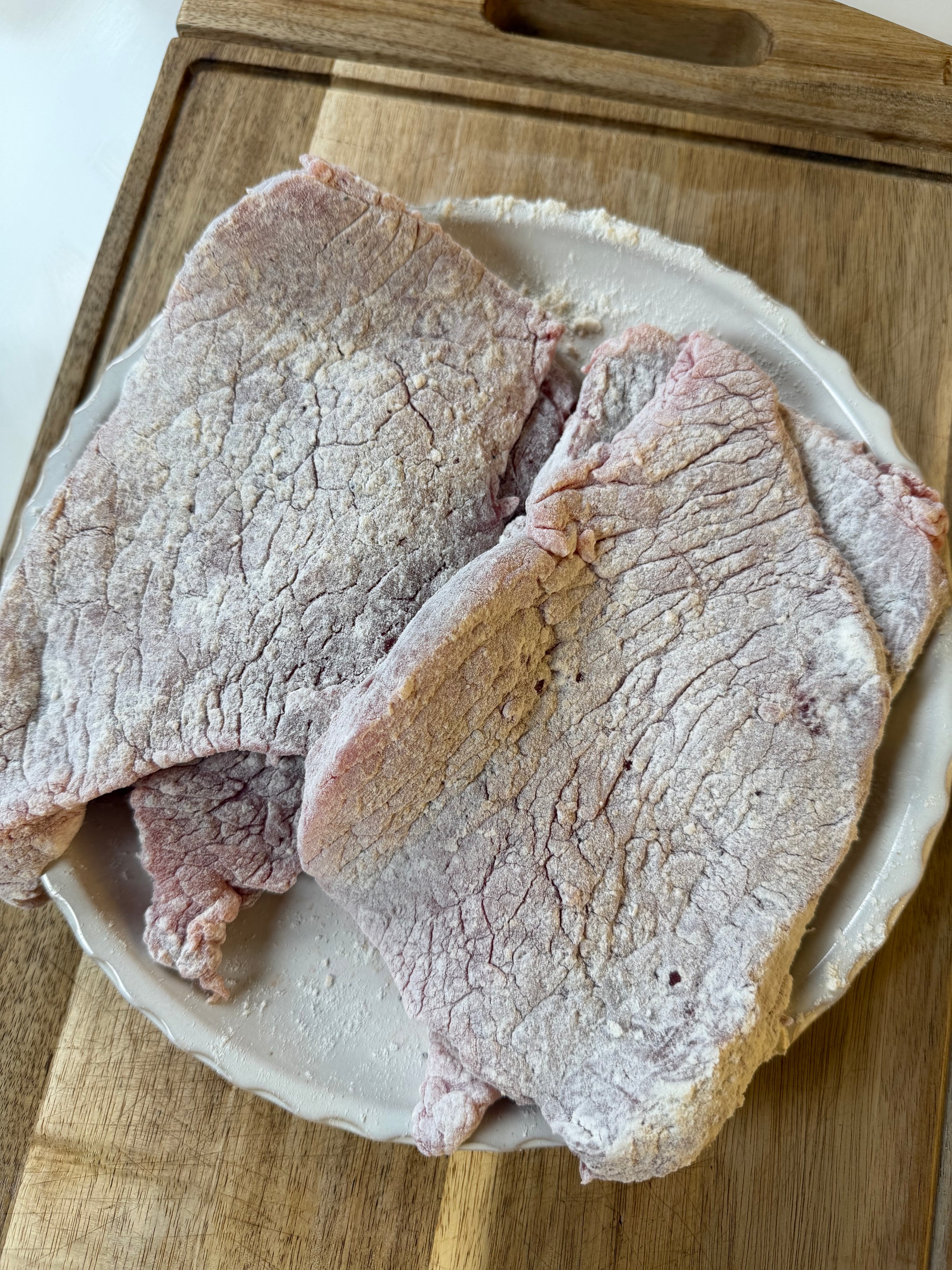 Two pieces of raw, lightly floured meat lie on a small white plate, which is placed on a wooden cutting board.