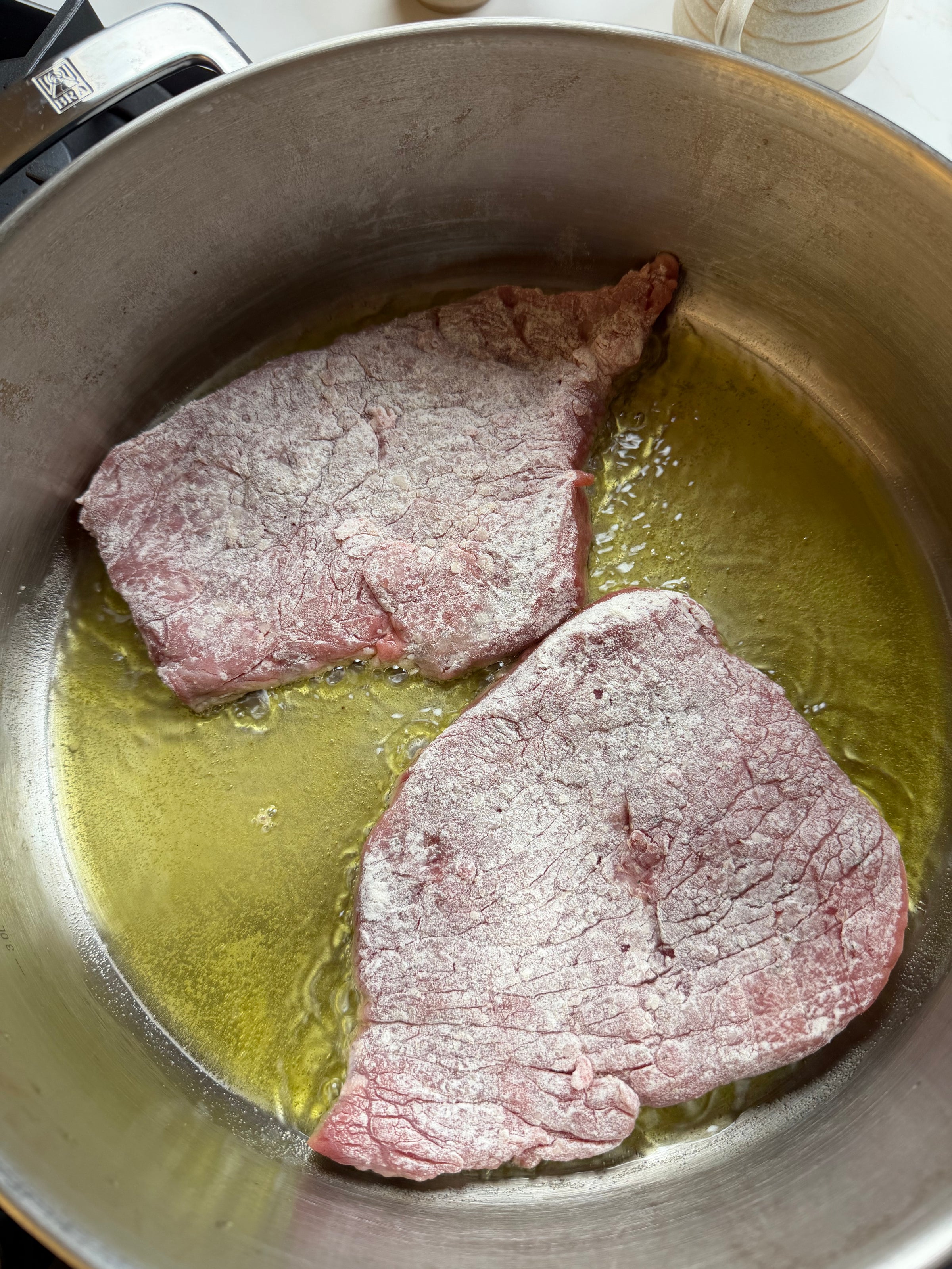 Two raw, flour-dusted beef steaks are lying in a stainless steel pan with hot oil, about to be seared.
