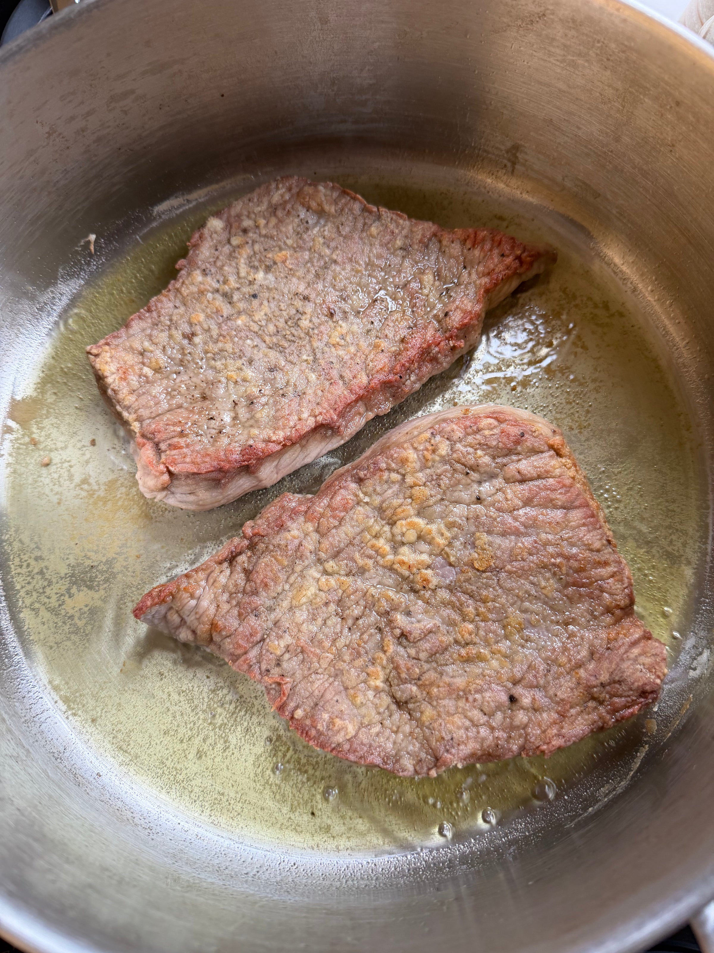 Two pieces of beef steak are being pan-fried in a metal pan with oil. The surfaces are browning and the oil is sizzling around the meat.