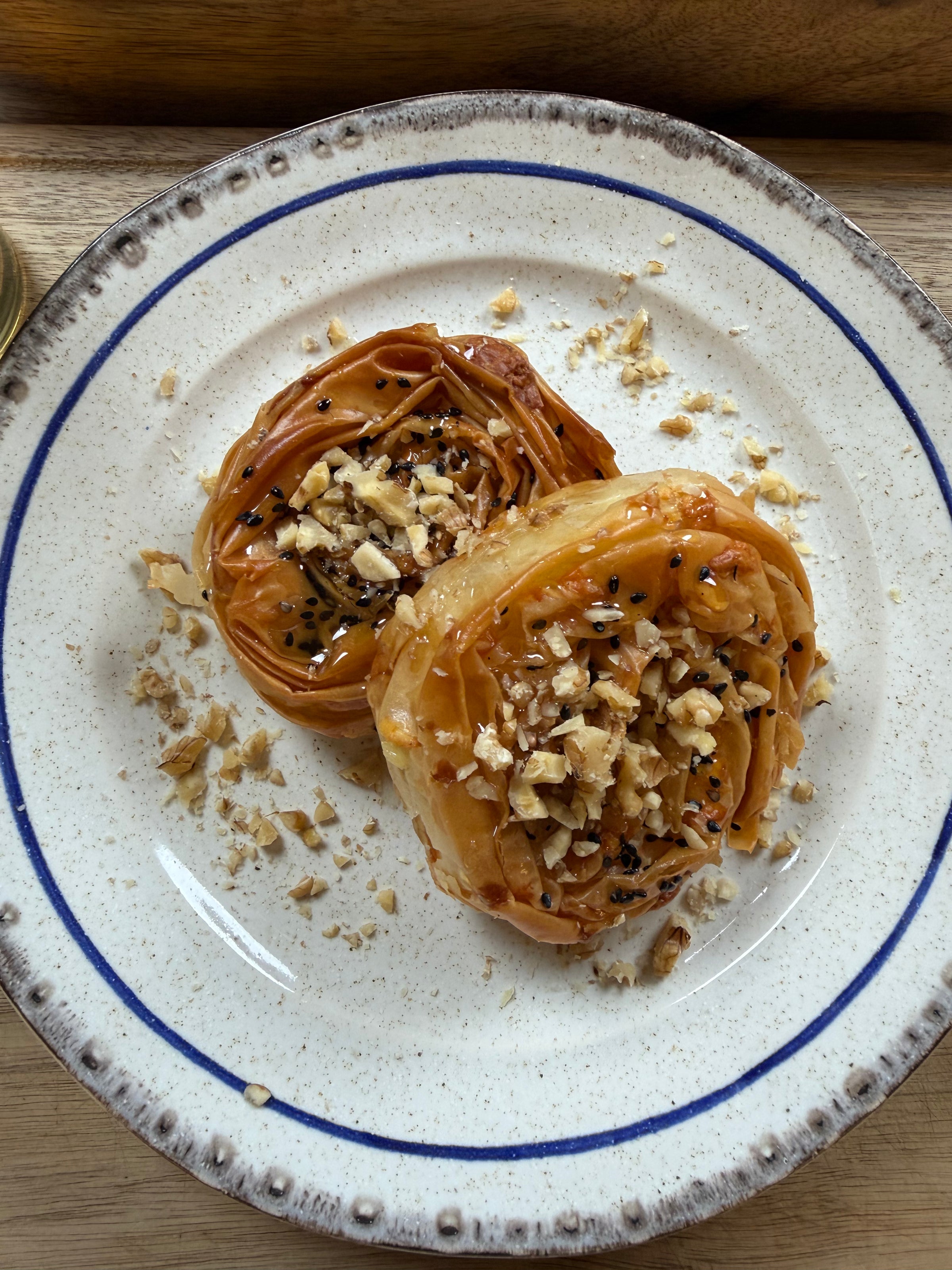 Two pieces of rolled pastry topped with chopped nuts and syrup are served on a speckled white plate with a blue rim, placed on a wooden surface.