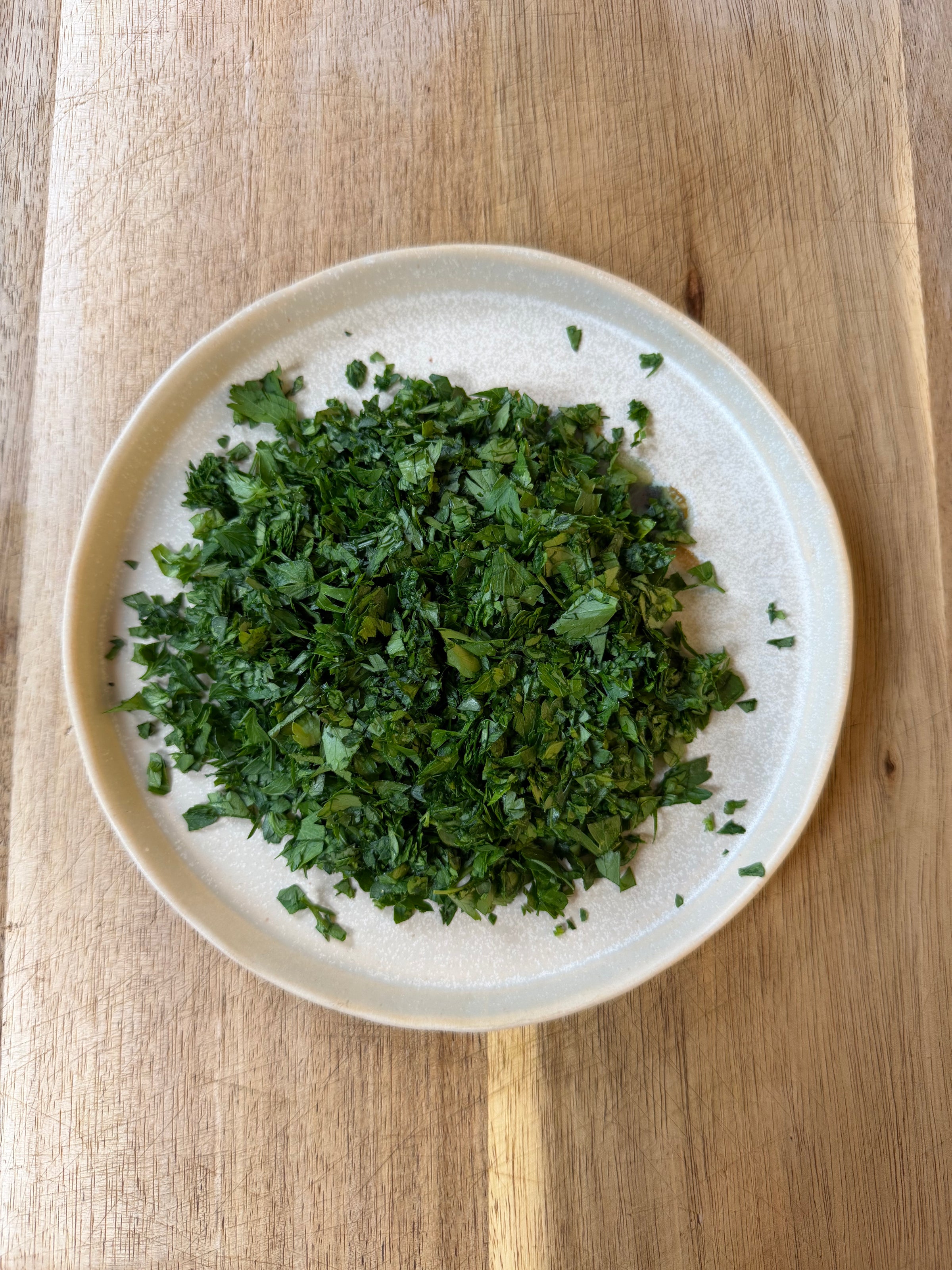 A white plate filled with finely chopped fresh parsley sits on a wooden surface.