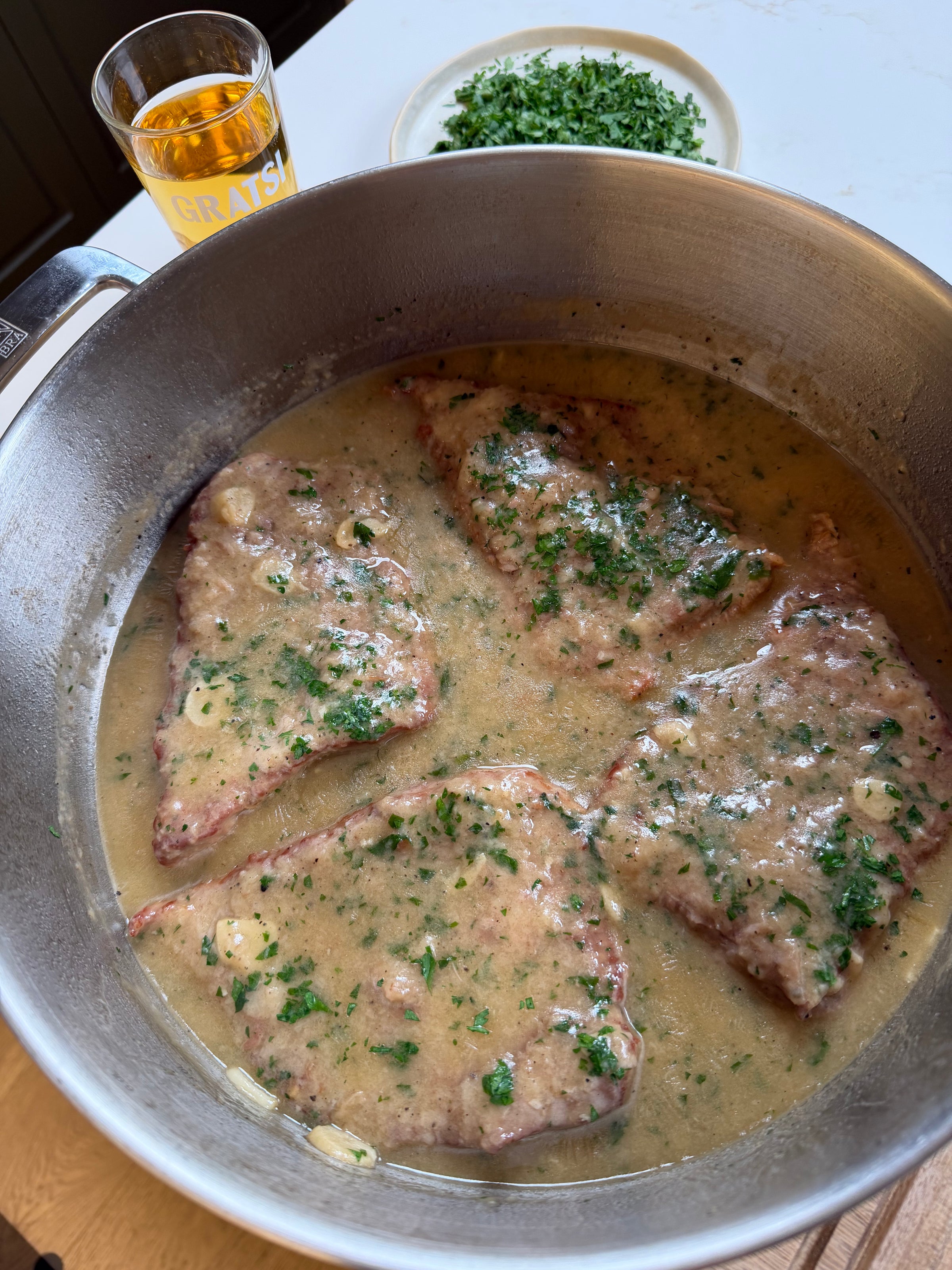 Four pieces of meat simmer in a creamy sauce with herbs in a stainless steel pan. A small bowl of chopped green herbs and a glass of amber liquid are in the background on a white surface.