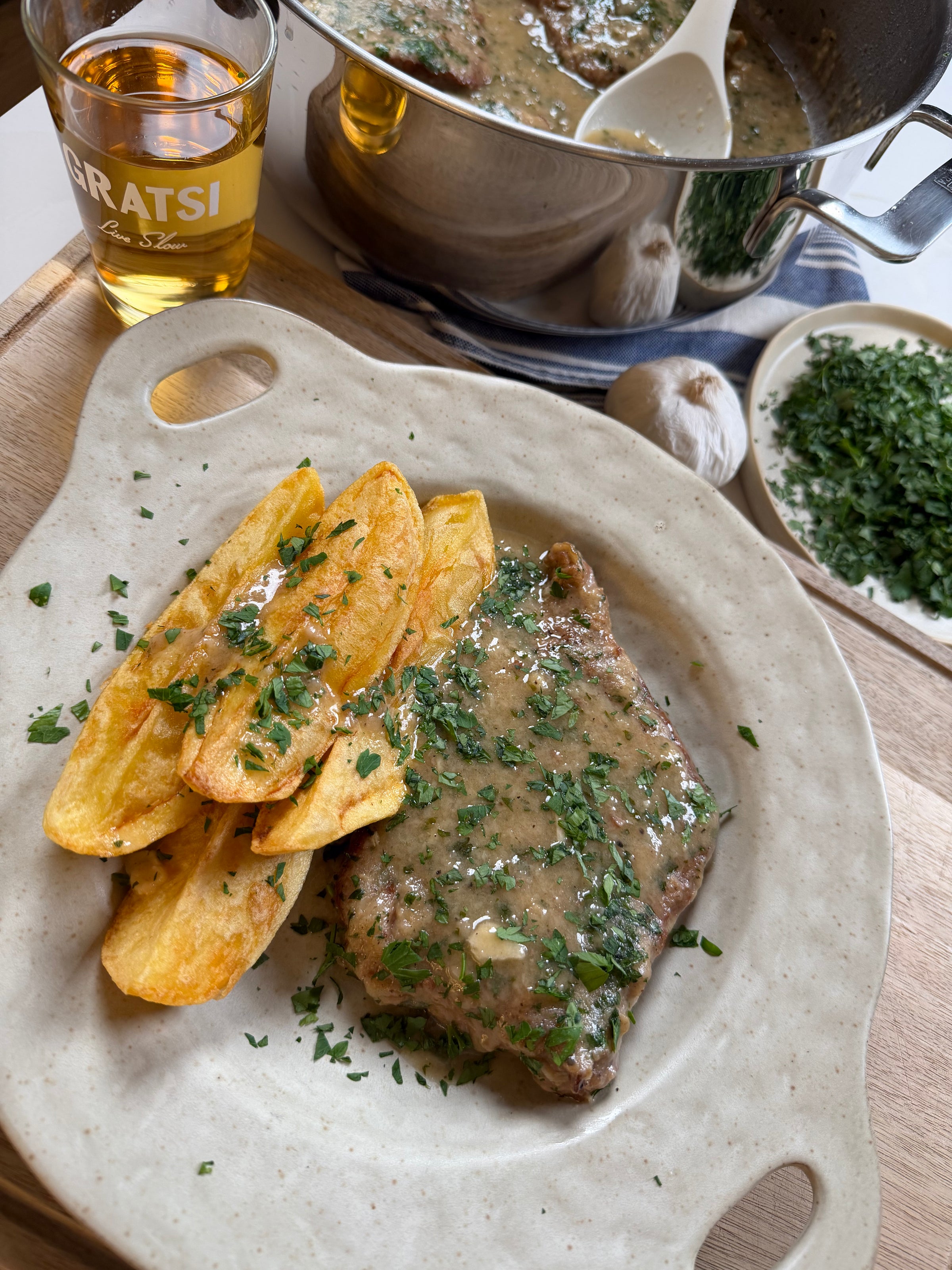 A plate with steak in creamy herb sauce, thick potato wedges, and chopped herbs. Nearby are a glass of drink, a pot of sauce with a spoon, garlic, and a bowl of herbs on a wooden table.