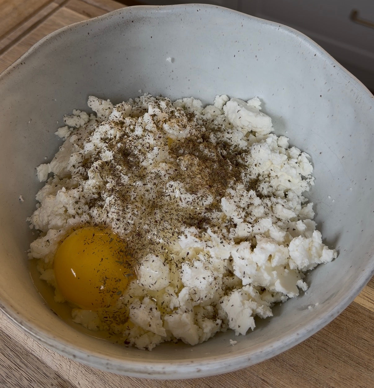 A white bowl contains crumbled ricotta cheese, a raw egg, and a sprinkling of black pepper, sitting on a wooden surface.