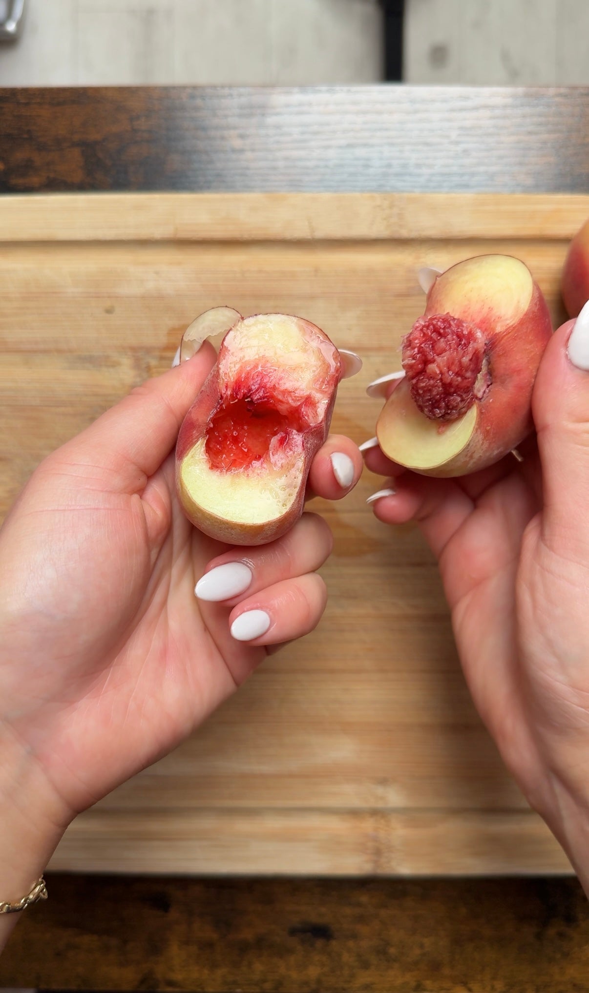 Two hands with manicured nails hold a halved peach over a wooden cutting board; one half has the pit, the other has a red, juicy center.