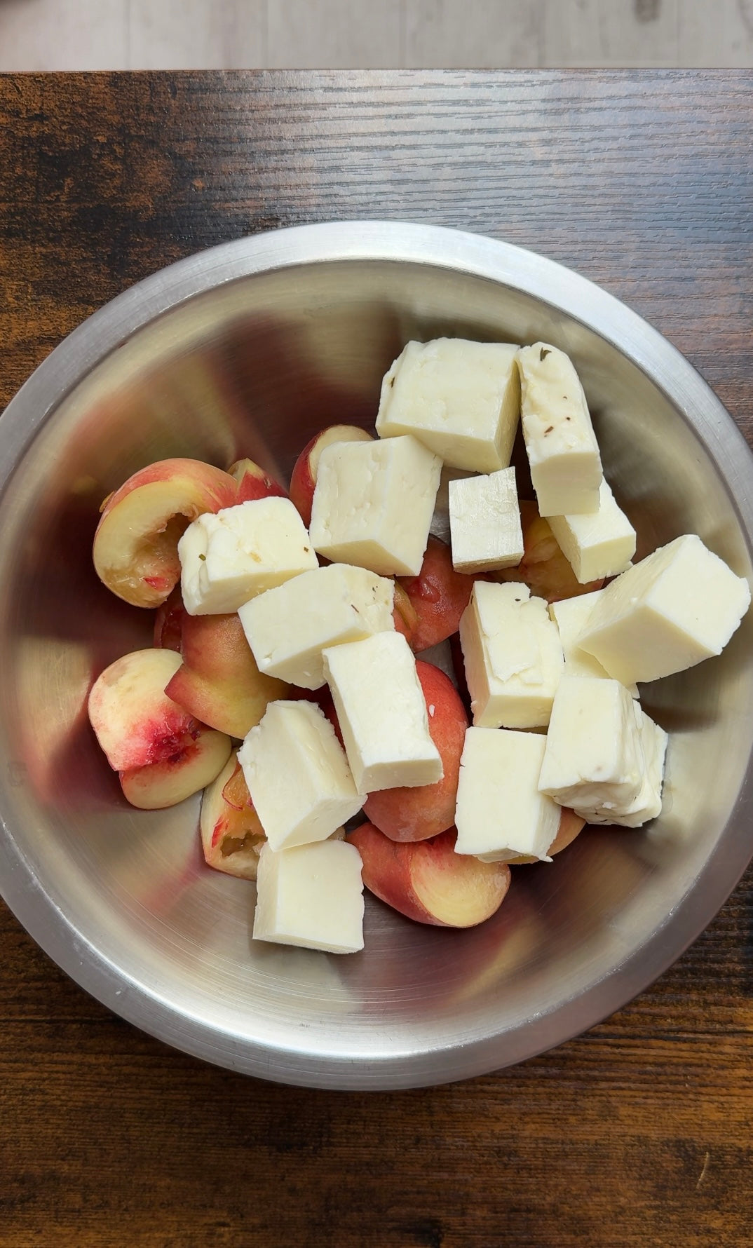 A metal bowl containing sliced peaches and large cubes of white cheese, placed on a dark wooden surface.