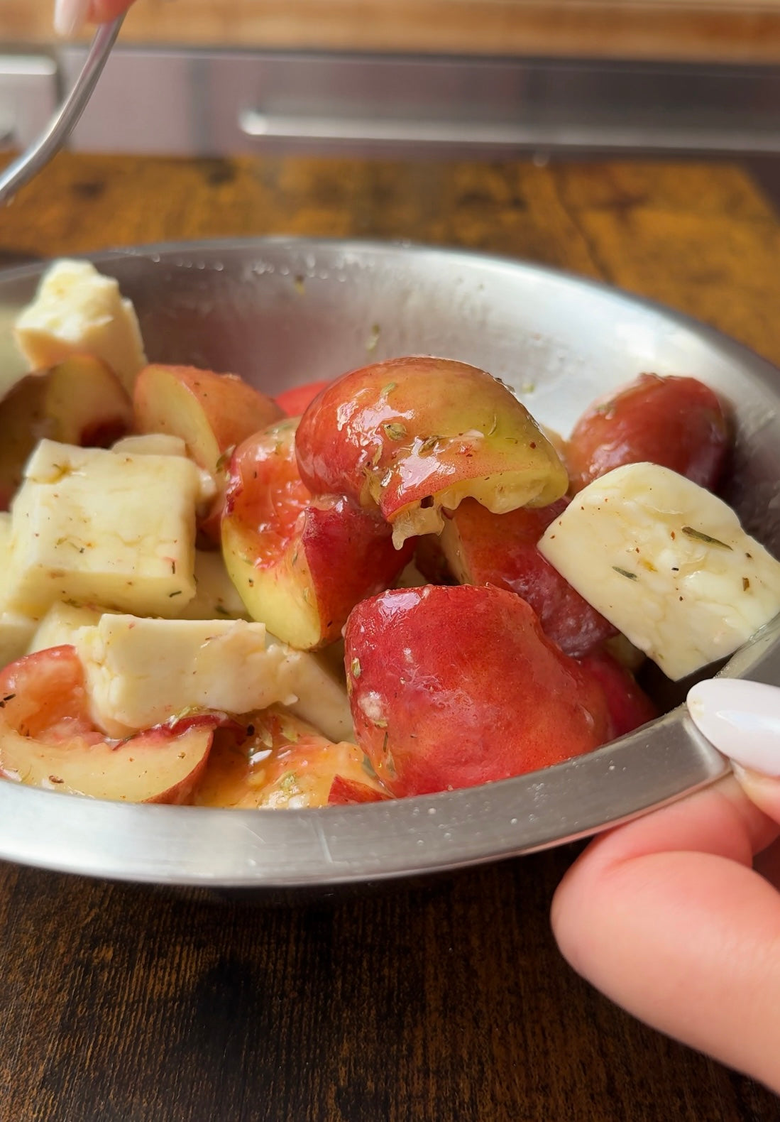 A close-up of a metal bowl containing sliced red potatoes and cubes of white cheese, garnished with herbs. A fork and a hand holding the bowl are visible.