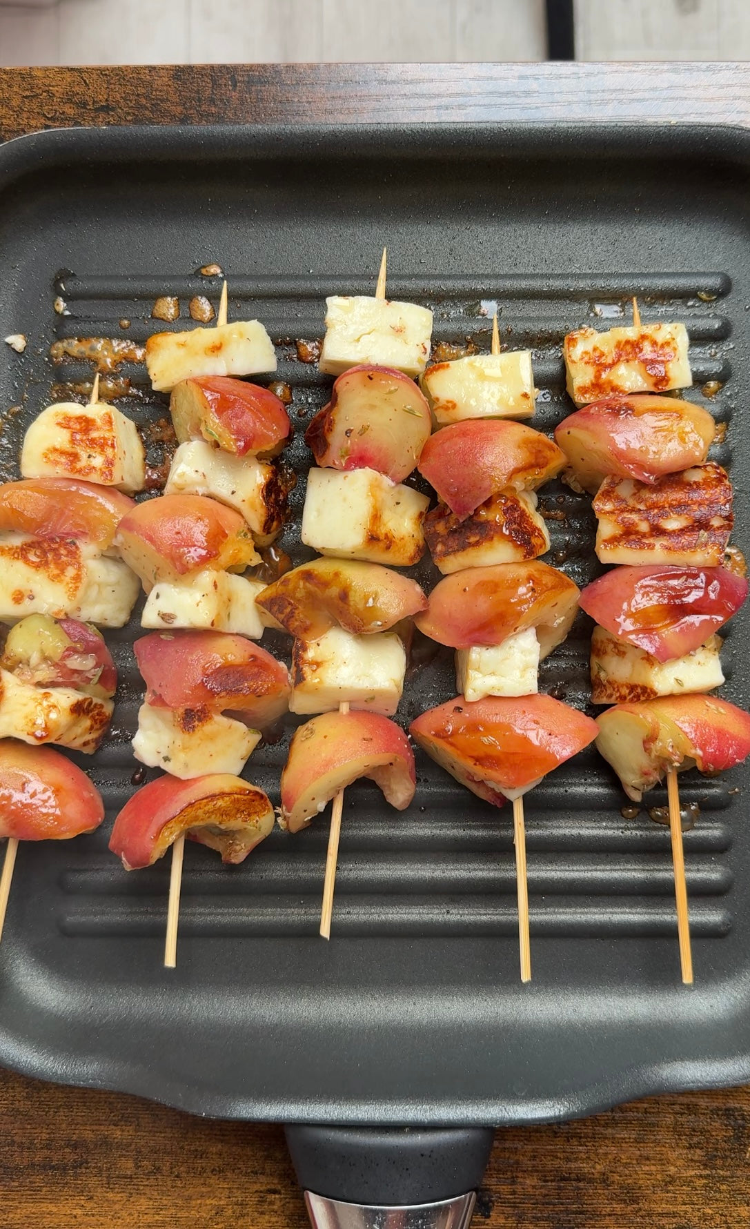 Skewers with grilled cheese cubes and sliced peaches are cooking on a ridged grill pan. The cheese is golden brown, and the peaches are slightly caramelized. The grill pan sits on a wooden surface.