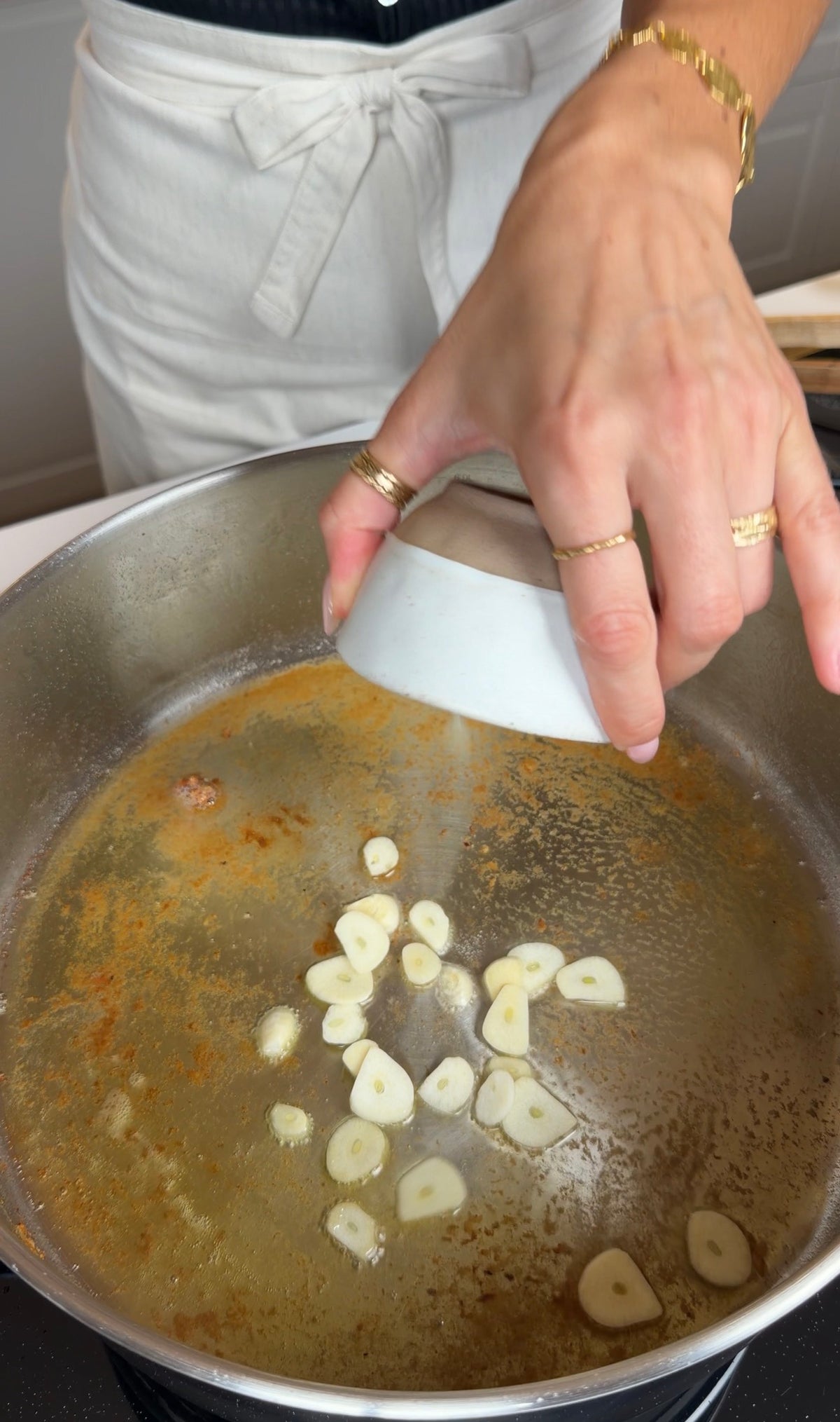 A person wearing rings adds sliced garlic from a small bowl into a large metal pan with oil on a stovetop.