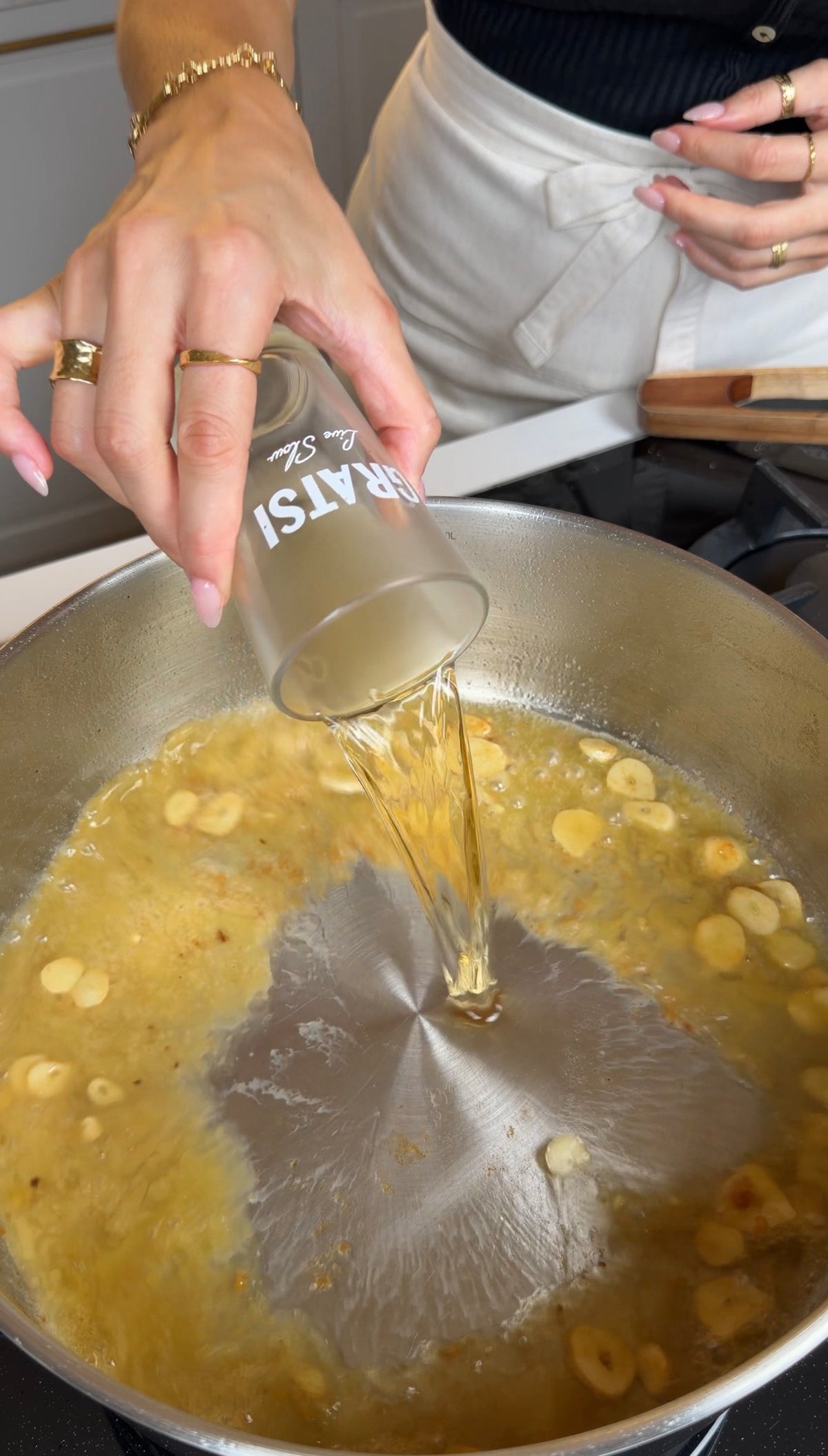 A person pours liquid from a glass into a skillet with sliced garlic simmering in oil on a stovetop.