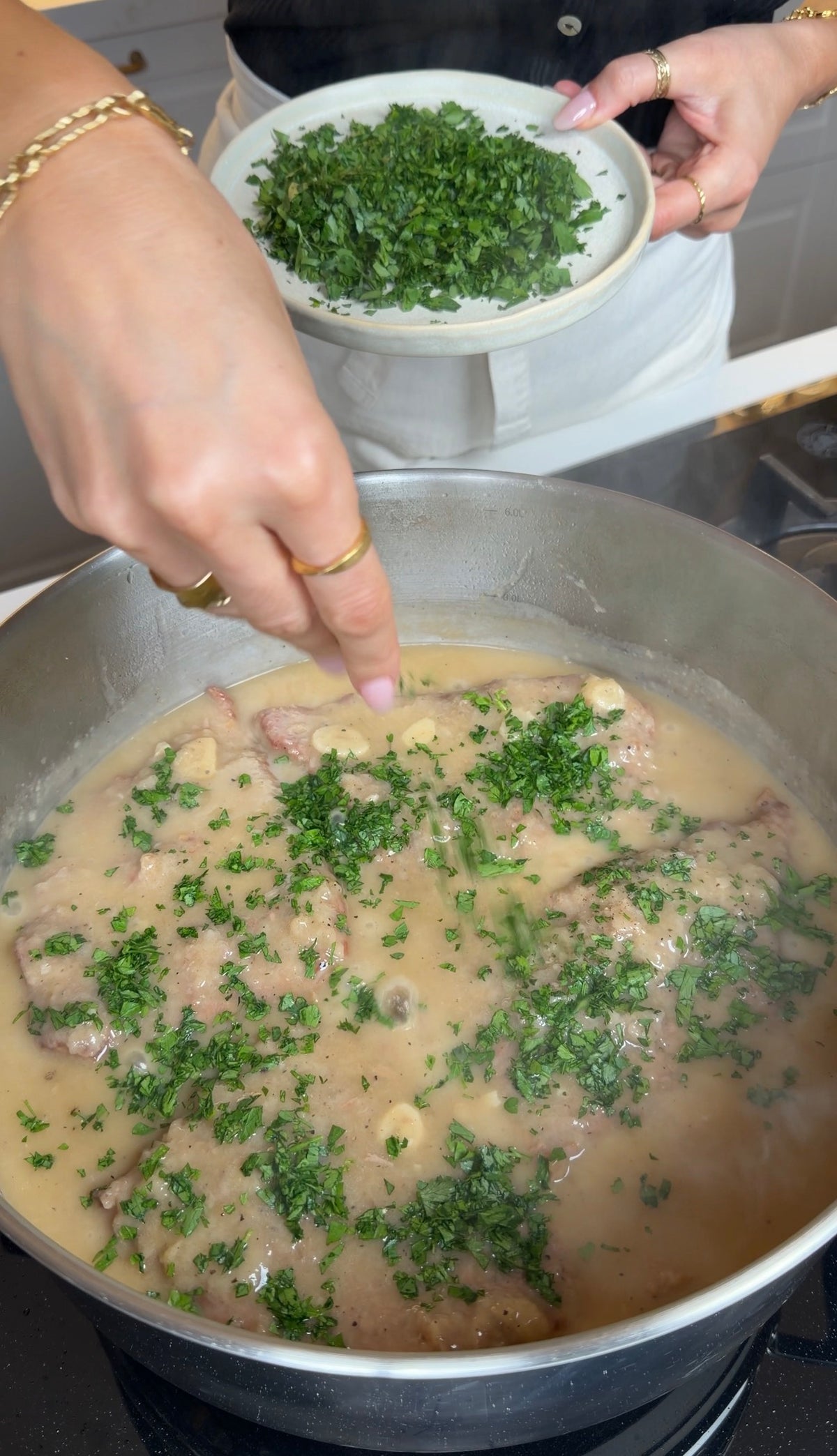A hand sprinkles fresh chopped parsley into a pan of meat simmering in a creamy sauce on the stove. A bowl of extra parsley is held in the other hand above the pan.