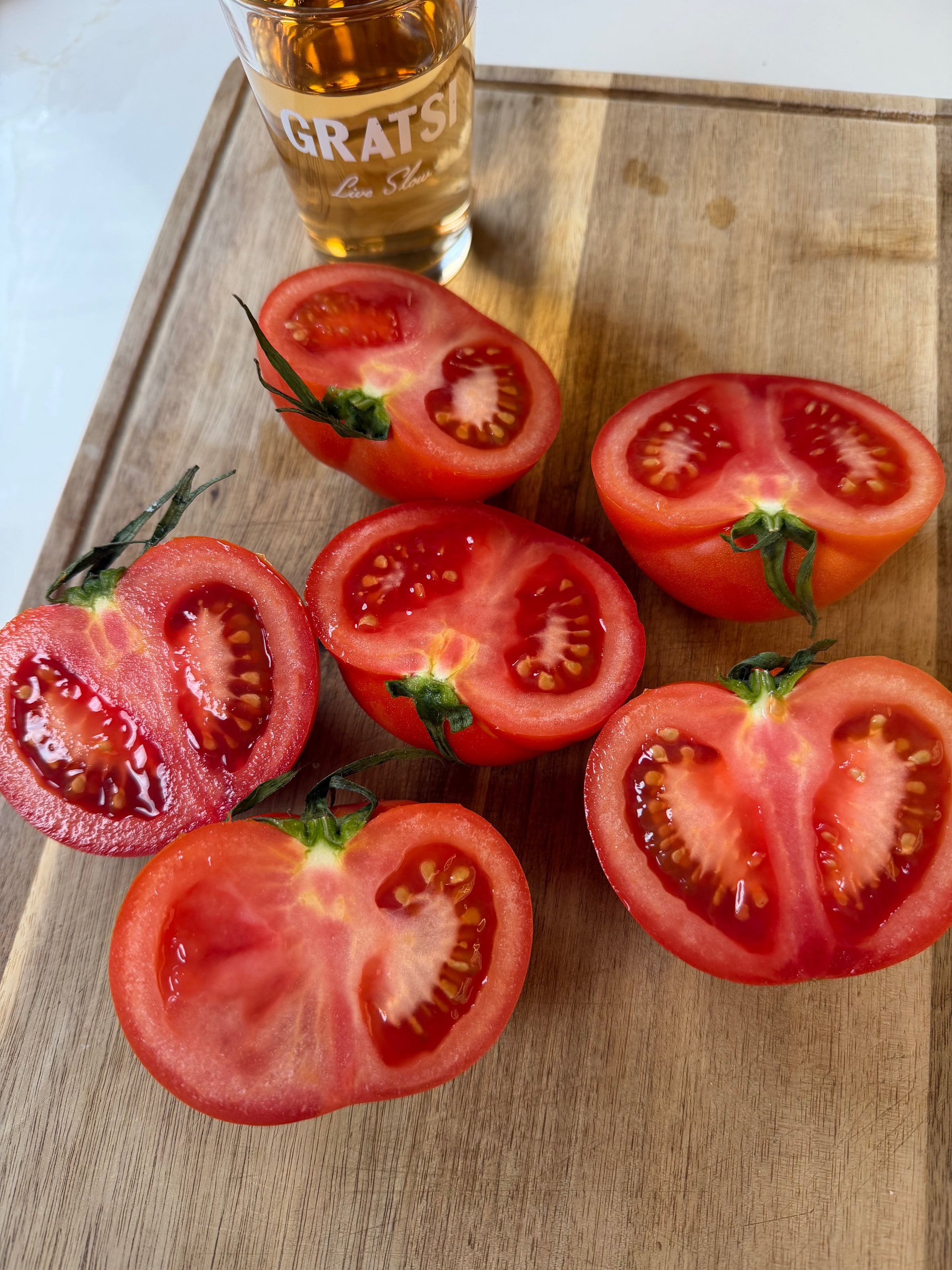Six ripe tomatoes cut in half are arranged on a wooden cutting board next to a glass with an amber drink, partially showing the word GRATSI on the glass.