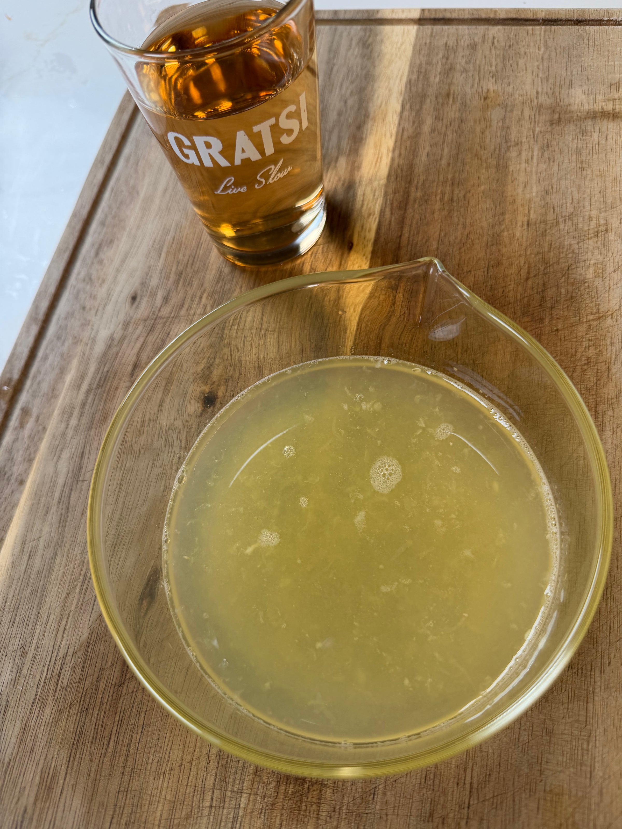 A glass bowl with yellowish liquid sits on a wooden surface next to a glass filled with amber-colored liquid labeled “GRATSI Live Slow”.
