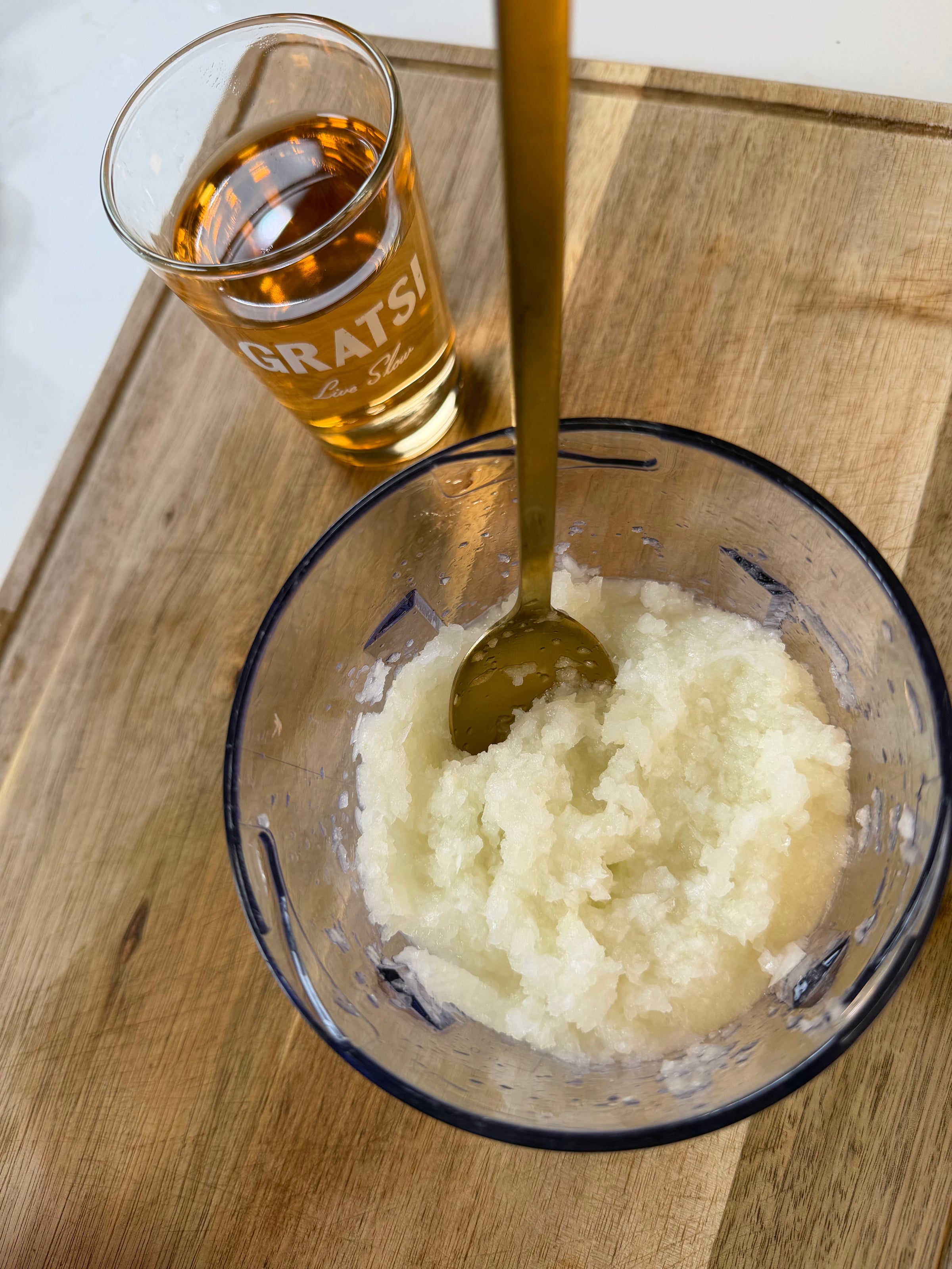A glass of amber liquid and a small bowl of finely grated or blended onion with a gold spoon rest on a wooden surface. Sunlight streams onto the scene.