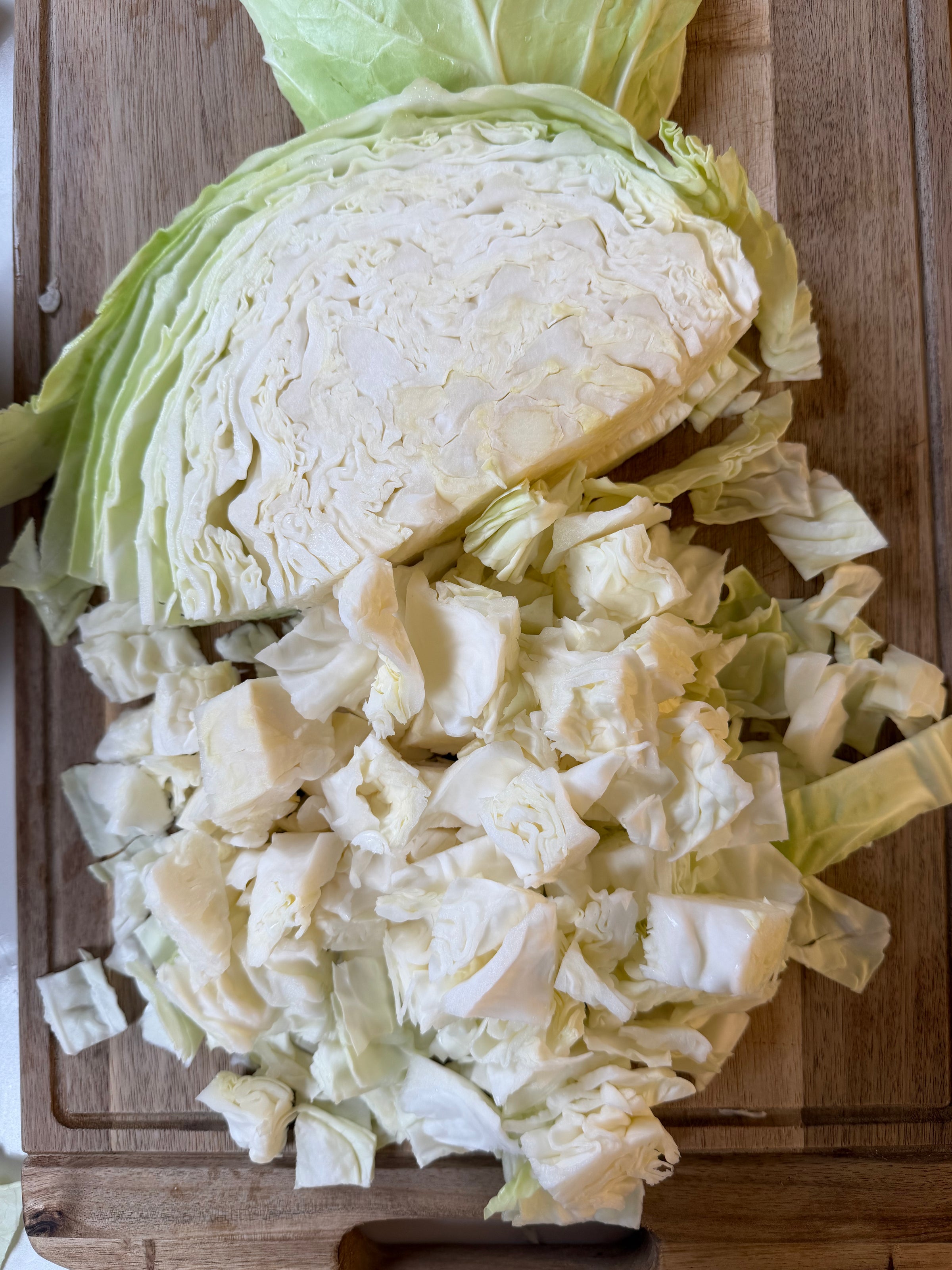 A halved green cabbage sits on a wooden cutting board, with one half chopped into small, square pieces.