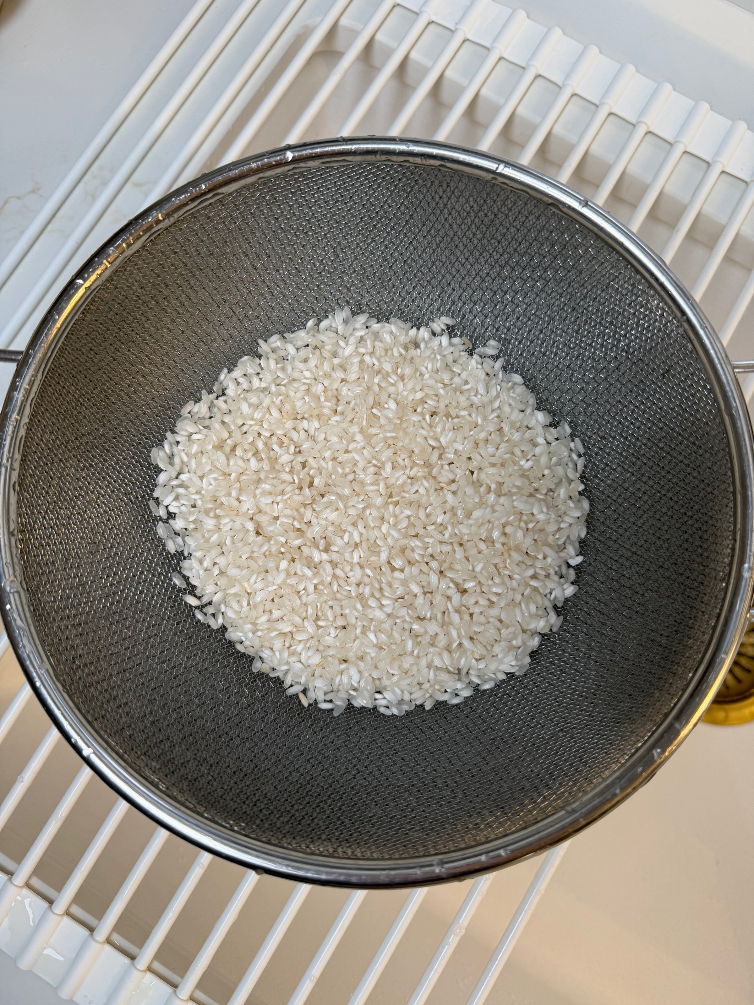 A metal strainer with uncooked white rice inside is placed on top of a white wire rack, with a countertop visible in the background.