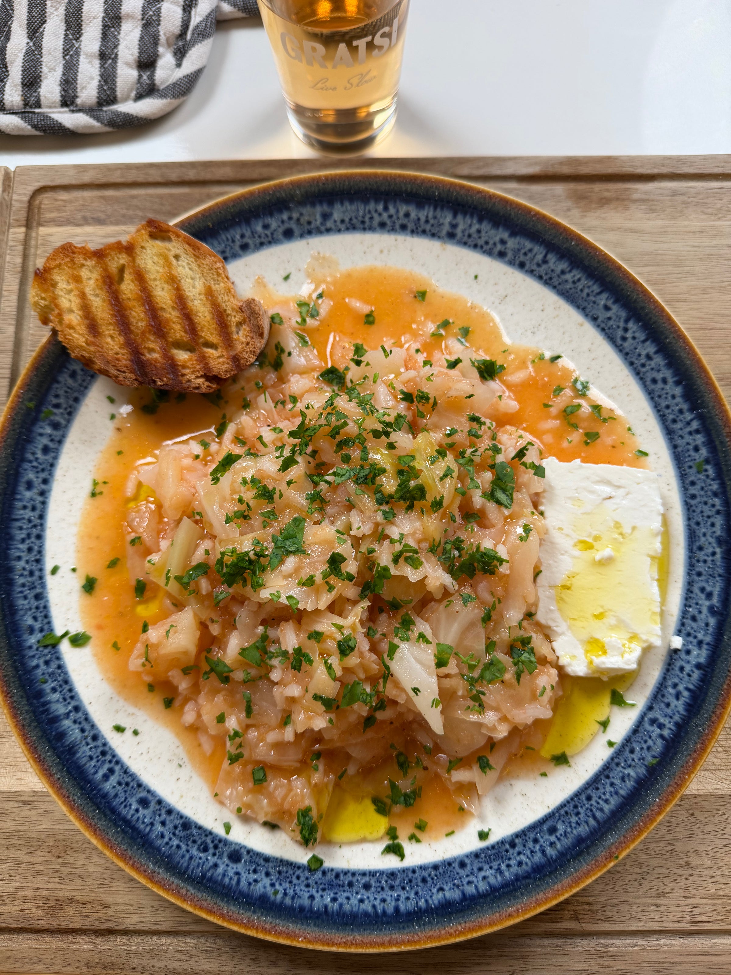 A plate of risotto topped with parsley, a slice of white cheese, and a drizzle of olive oil, served with a piece of grilled bread. A glass of golden beverage sits in the background on a wooden tray.