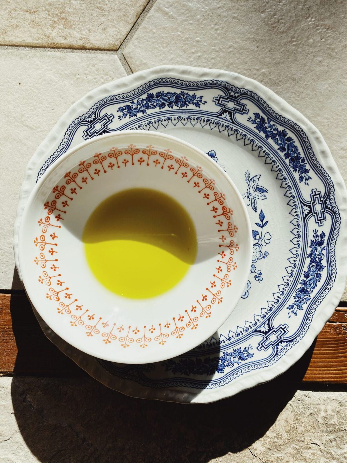 A small bowl with orange decorative patterns filled with yellow olive oil sits on a larger blue and white patterned plate, both placed on a tiled surface in sunlight.