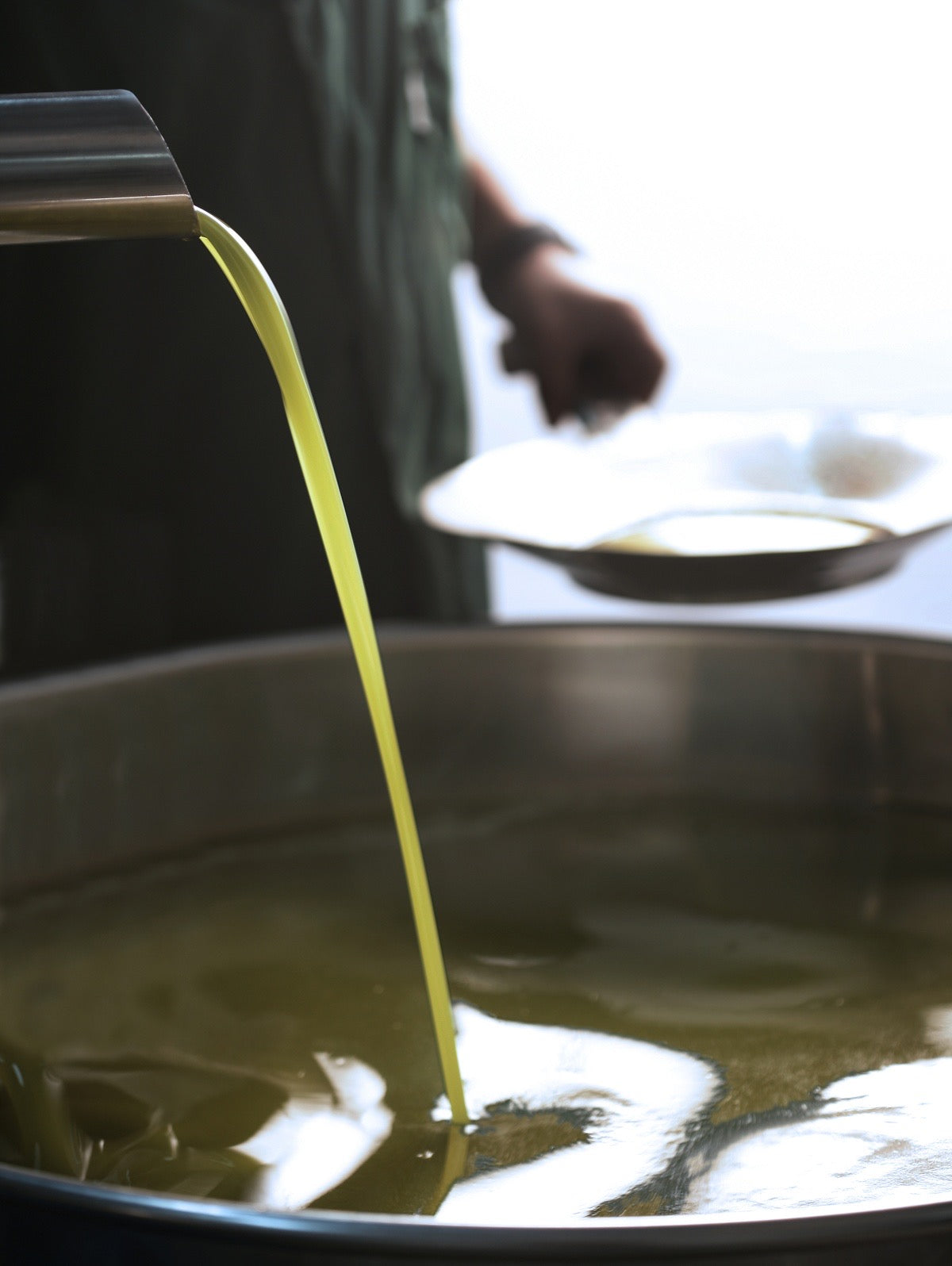 A stream of yellowish-green liquid is being poured from a metal spout into a large stainless steel container, with a person holding a plate in the blurred background.
