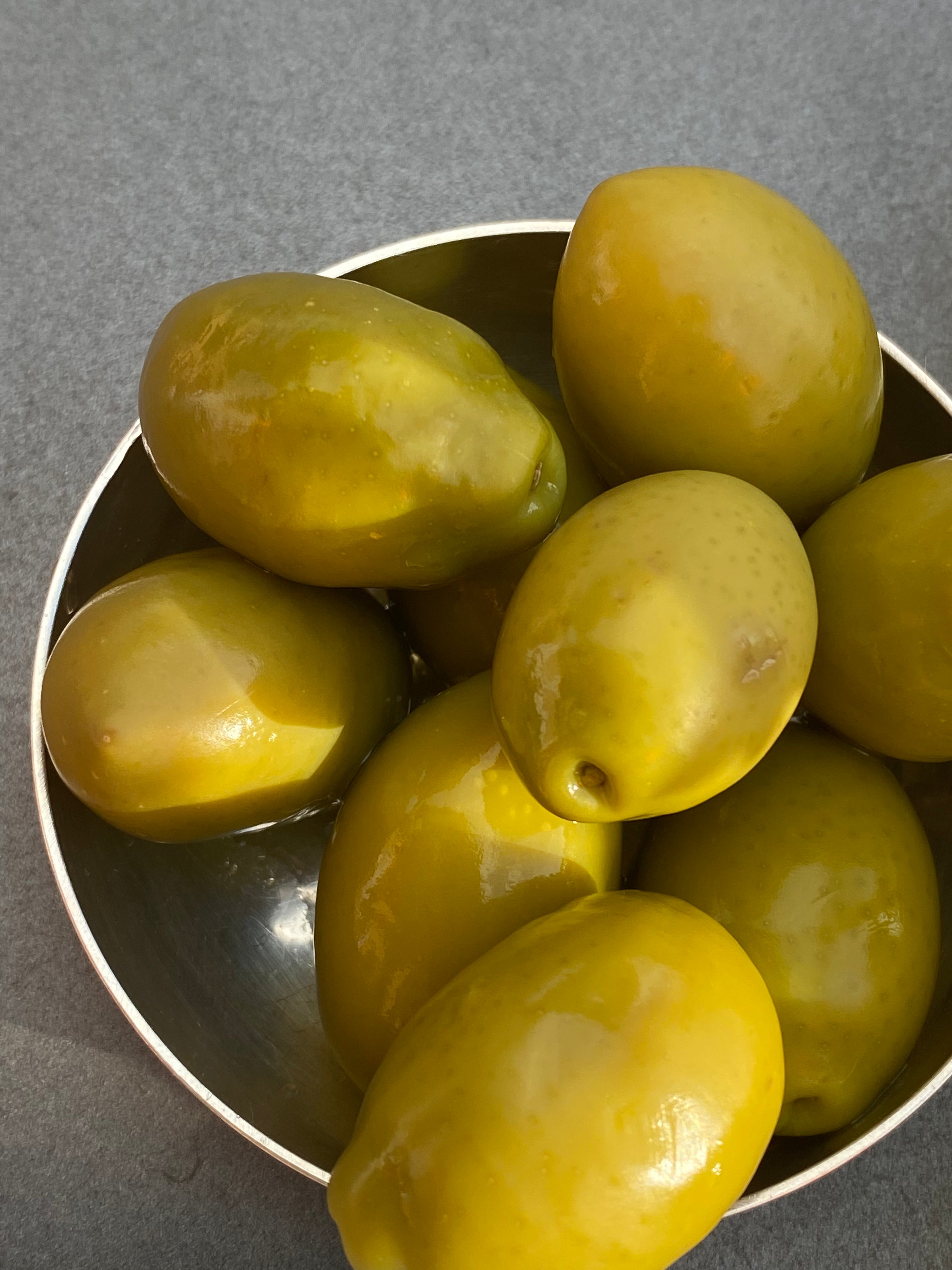 A close-up of a metal bowl filled with large, green olives on a gray surface.