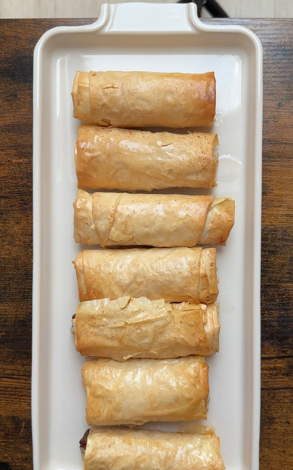 Six golden-brown, phyllo-wrapped pastry rolls are neatly arranged in a white rectangular dish on a wooden table.