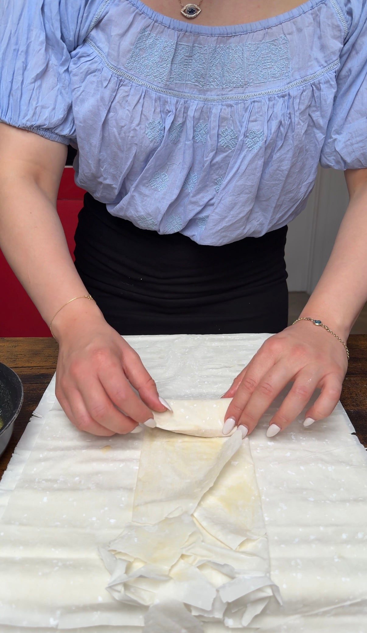 A person folds a sheet of pastry on a wooden table. There is a pan and a bowl nearby. The person wears a blue top and black skirt, and only their torso and hands are visible.