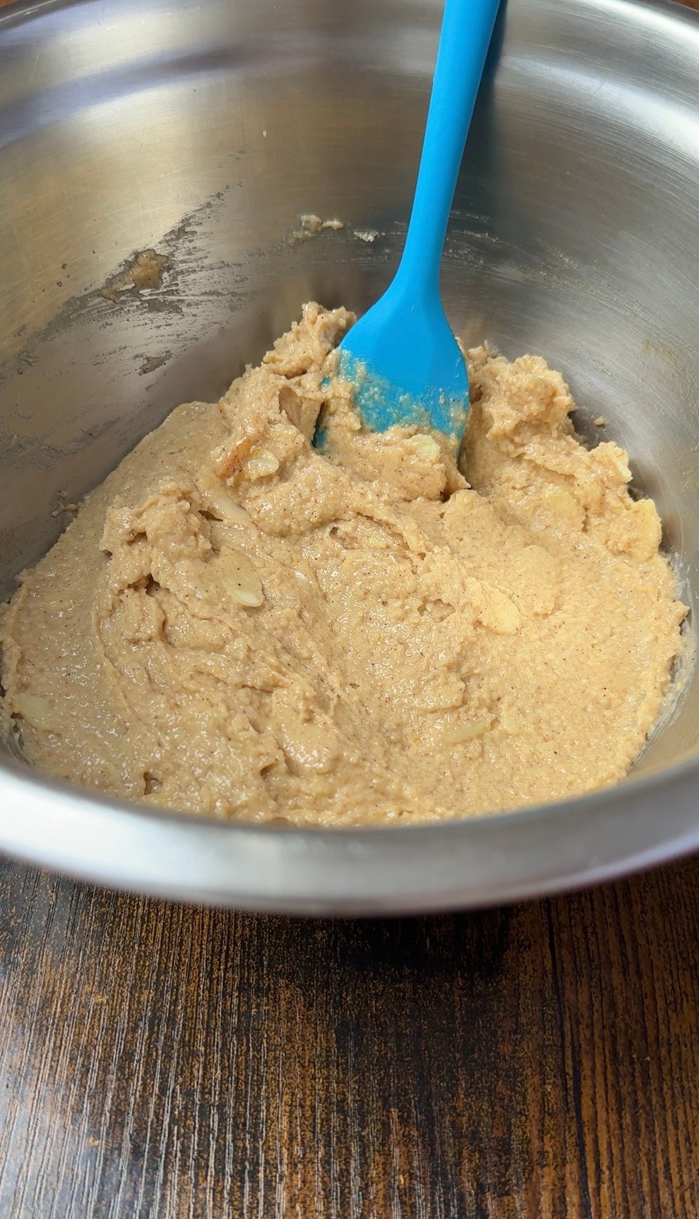 A blue spatula in a metal bowl with light brown cookie dough being mixed, sitting on a wooden surface.