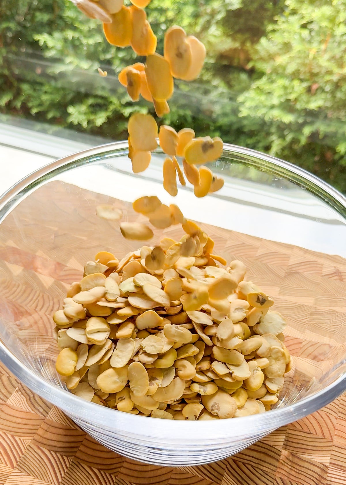 Slices of dried yellow beans are being poured into a glass bowl, which sits on a patterned wooden surface. Green foliage is visible outside the window in the background.