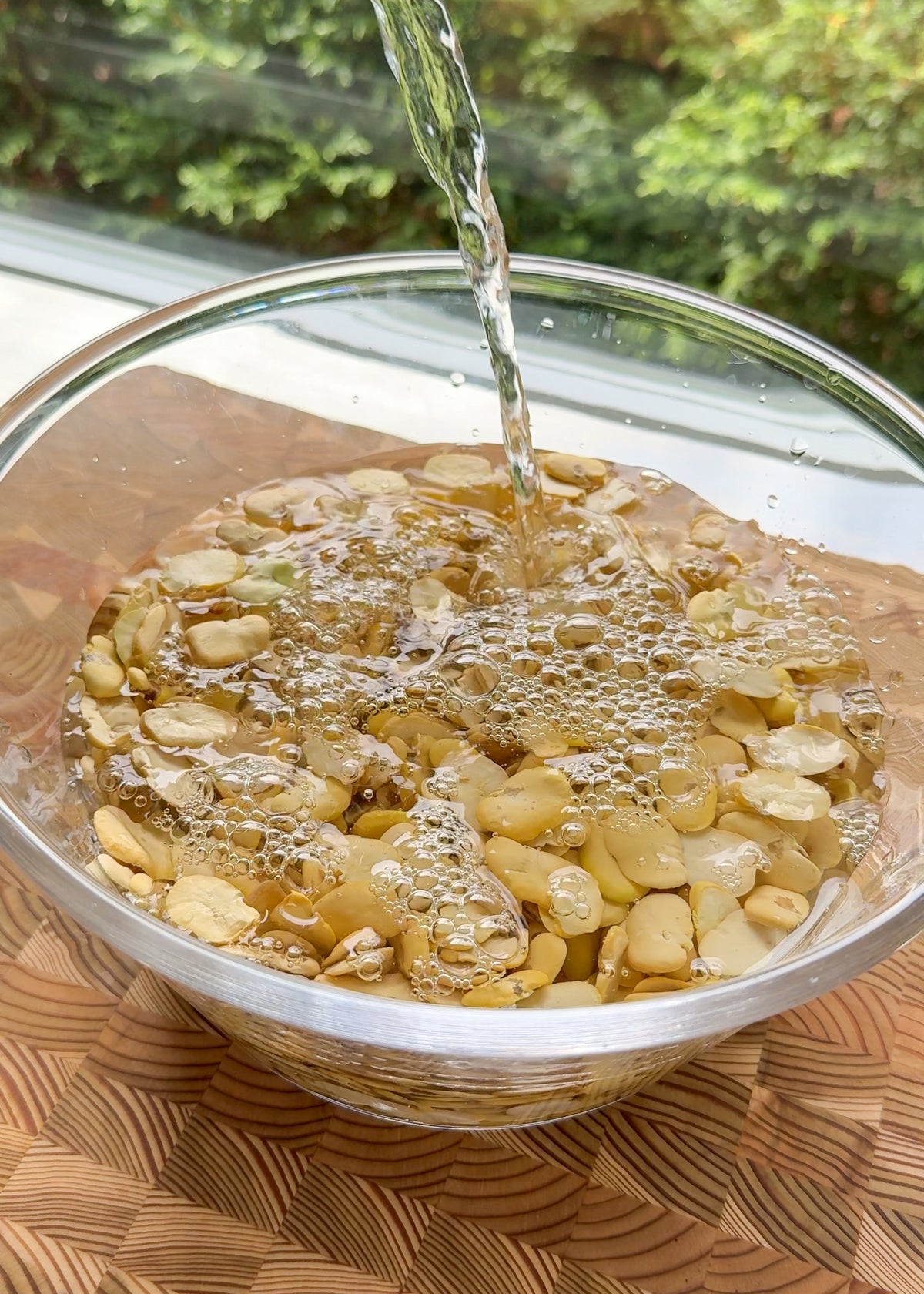 Water is being poured into a glass bowl filled with sliced almonds, causing bubbles to form on the surface. The bowl sits on a wooden surface near a window with greenery outside.