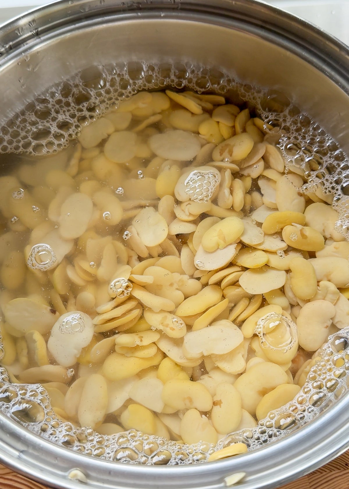 A stainless steel pot filled with sliced lima beans soaking in water, with bubbles forming on the surface.