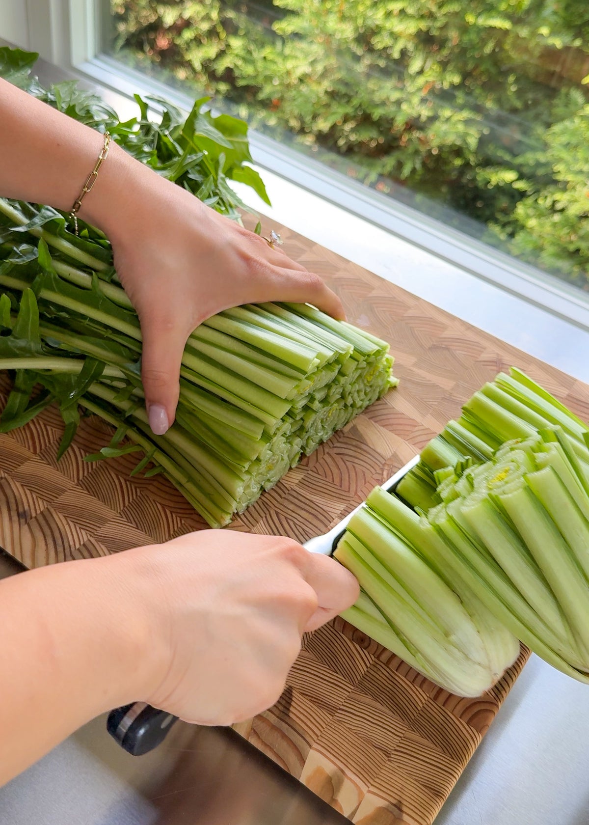A person uses a knife to slice a large bunch of green celery stalks on a wooden cutting board near a window with greenery outside.