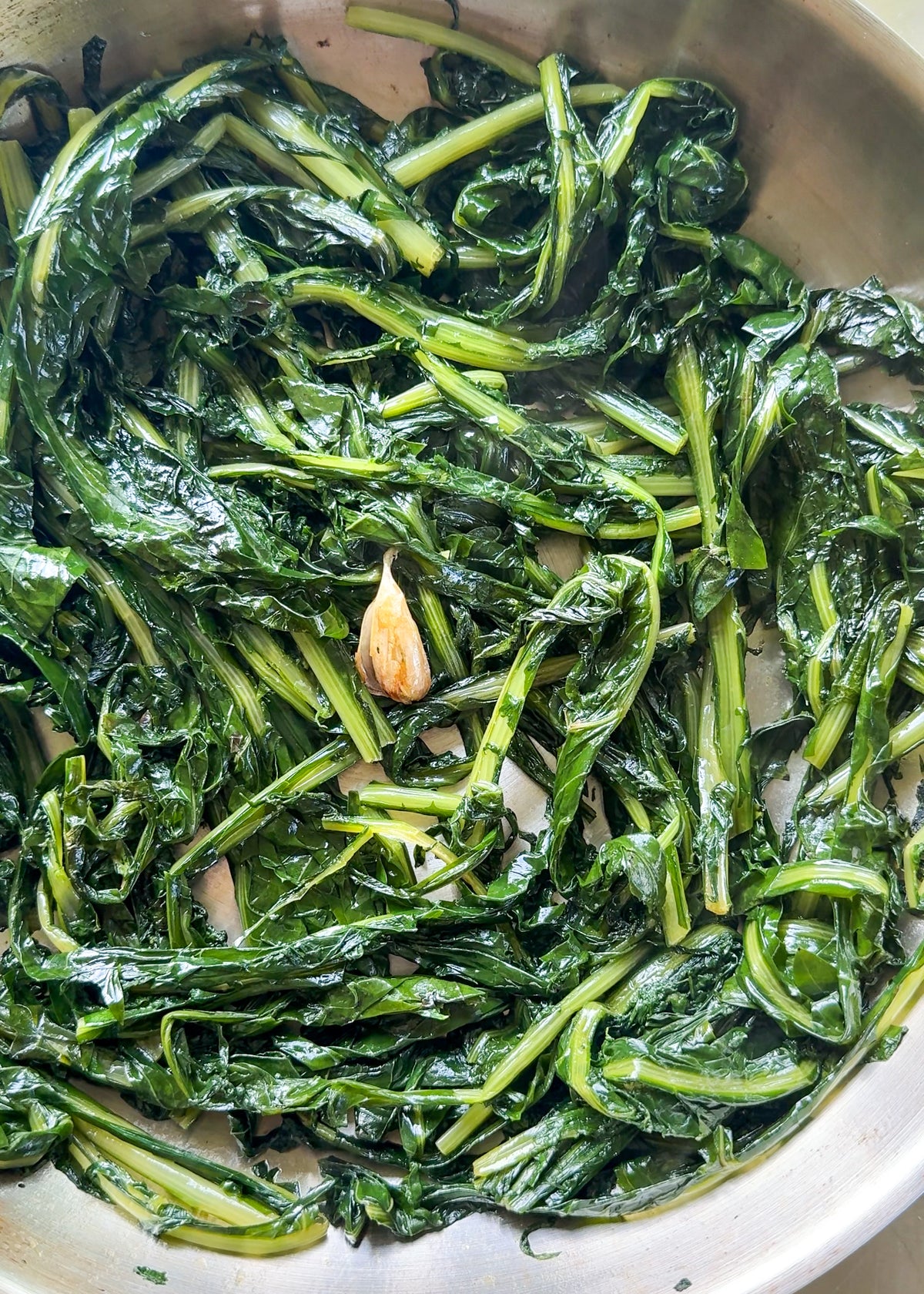 Sautéed leafy greens with a single clove of browned garlic in a stainless steel pan.