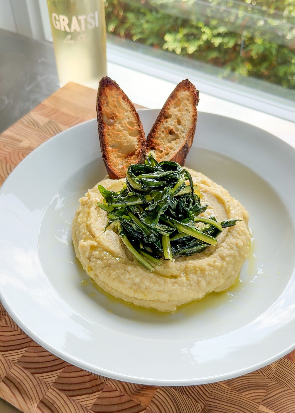 A white plate with creamy hummus topped with sautéed greens, served with two toasted bread slices. A bottle labeled Gratsi and greenery outside a window are visible in the background.