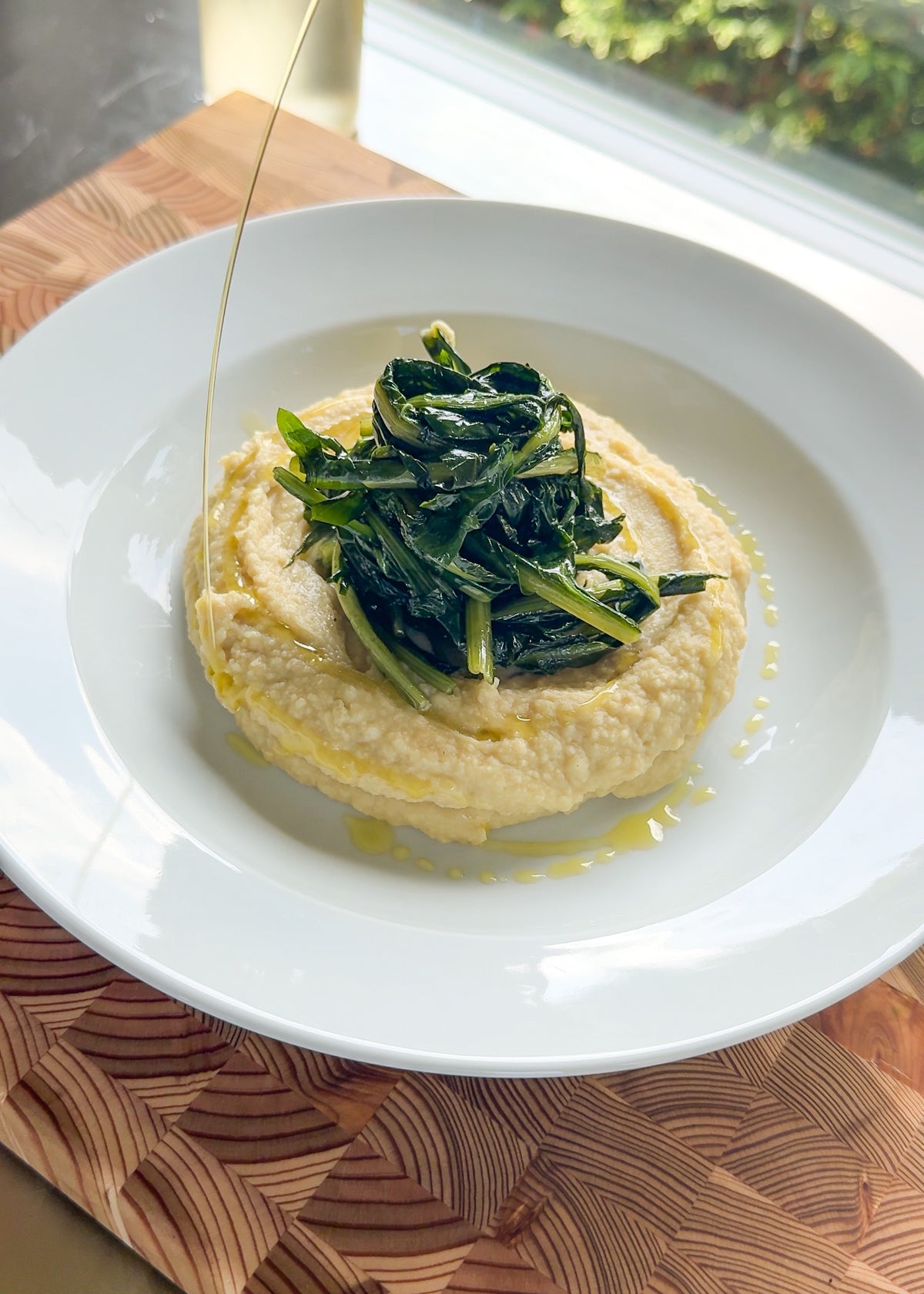 A white plate with creamy polenta topped with sautéed greens, drizzled with olive oil. The plate sits on a wooden board near a window with greenery outside.