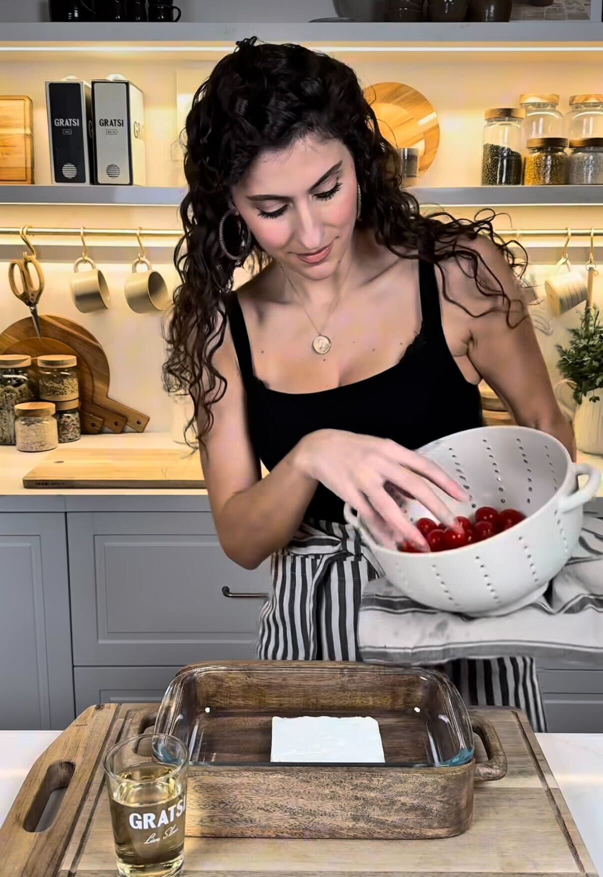 A woman in a kitchen stands at a counter, holding a white colander with cherry tomatoes. In front of her are a glass baking dish with a block of cheese, a glass of oil, and a wooden cutting board. Shelves with jars are in the background.