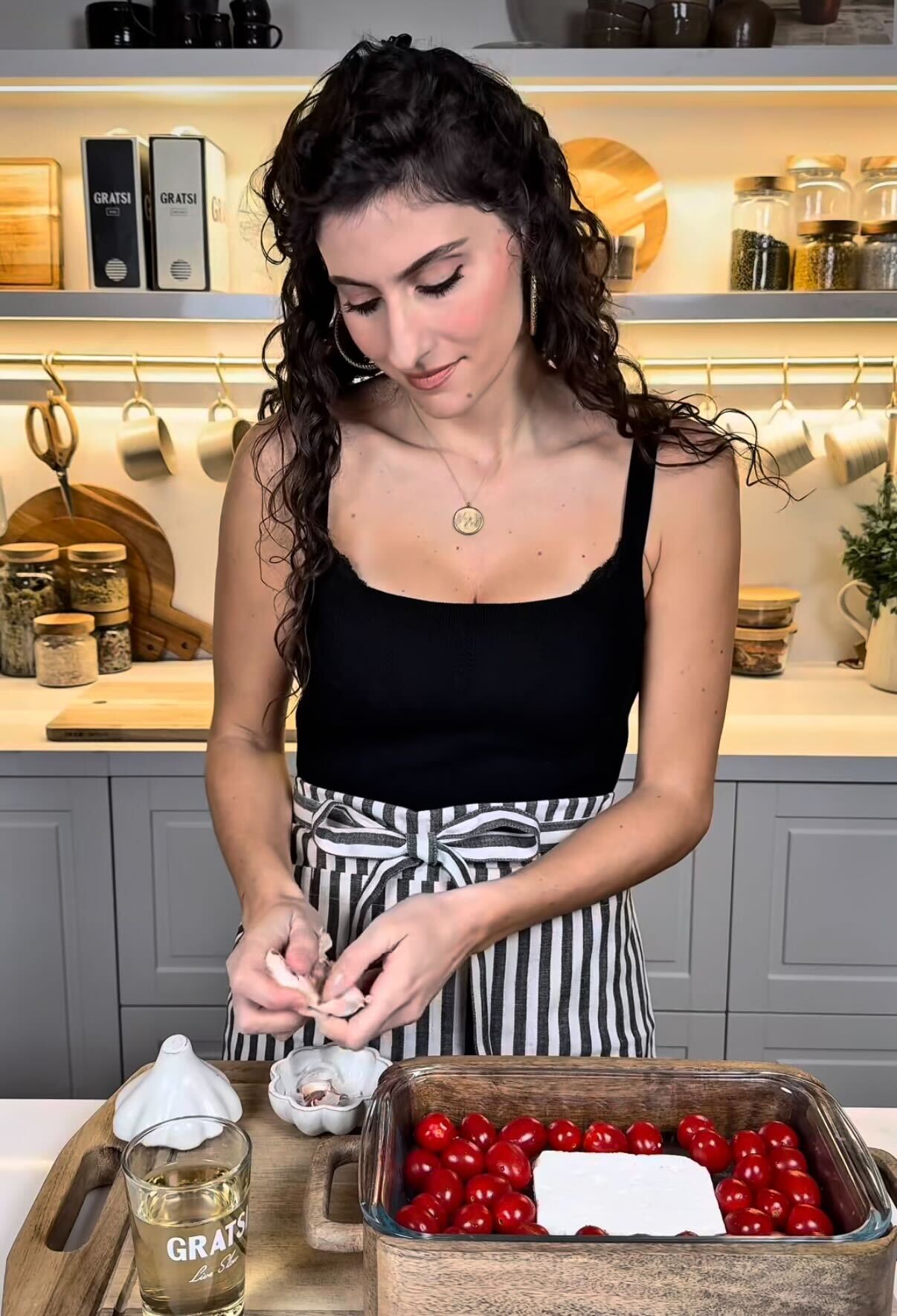 A woman in a striped apron prepares food in a modern kitchen, crumbling an ingredient over a baking dish filled with cherry tomatoes and a block of cheese. Various jars and kitchen items are visible on shelves behind her.