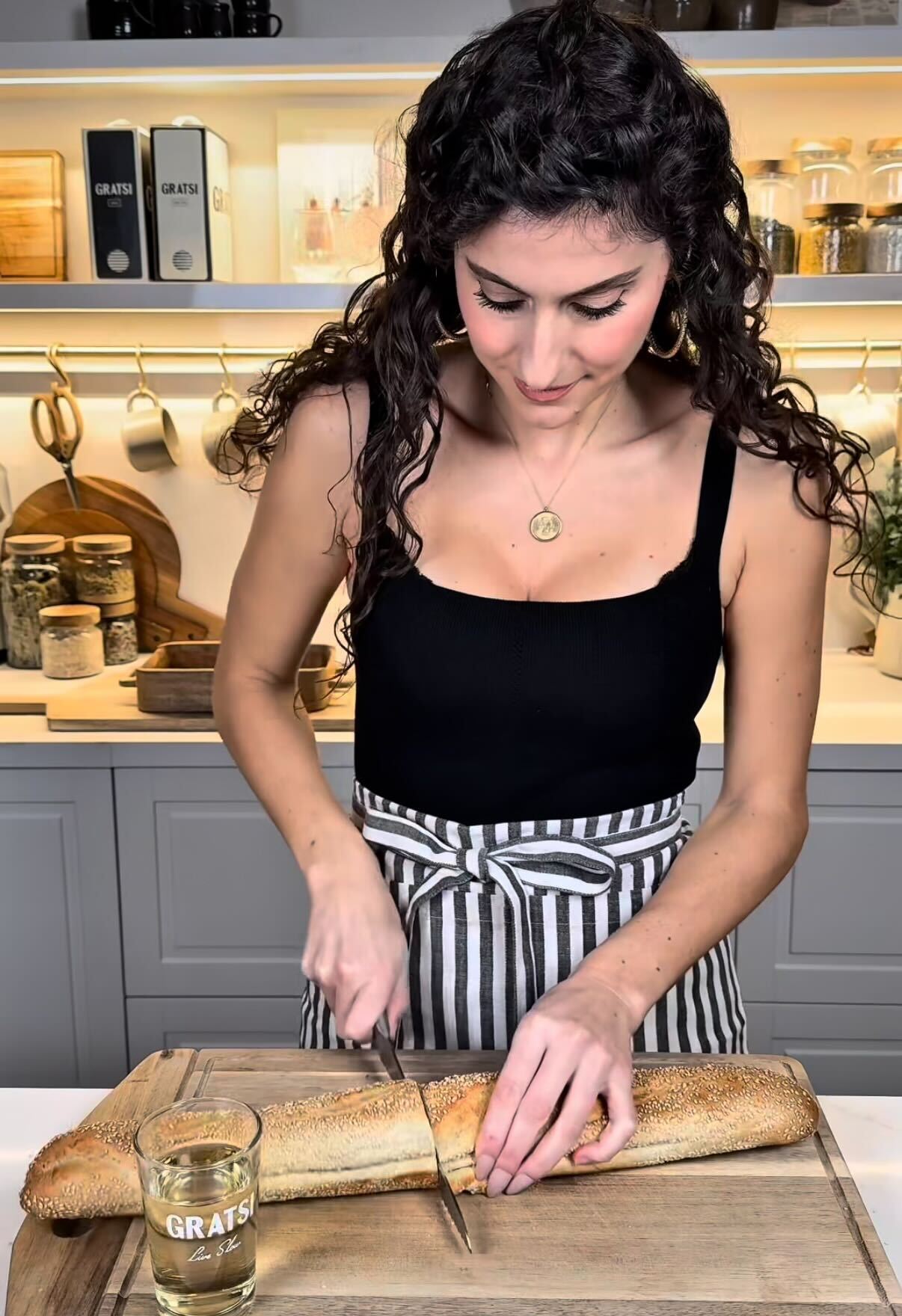 A woman with curly hair wearing a black top and striped apron slices a baguette on a wooden cutting board in a modern kitchen. A glass of white wine sits nearby on the counter.