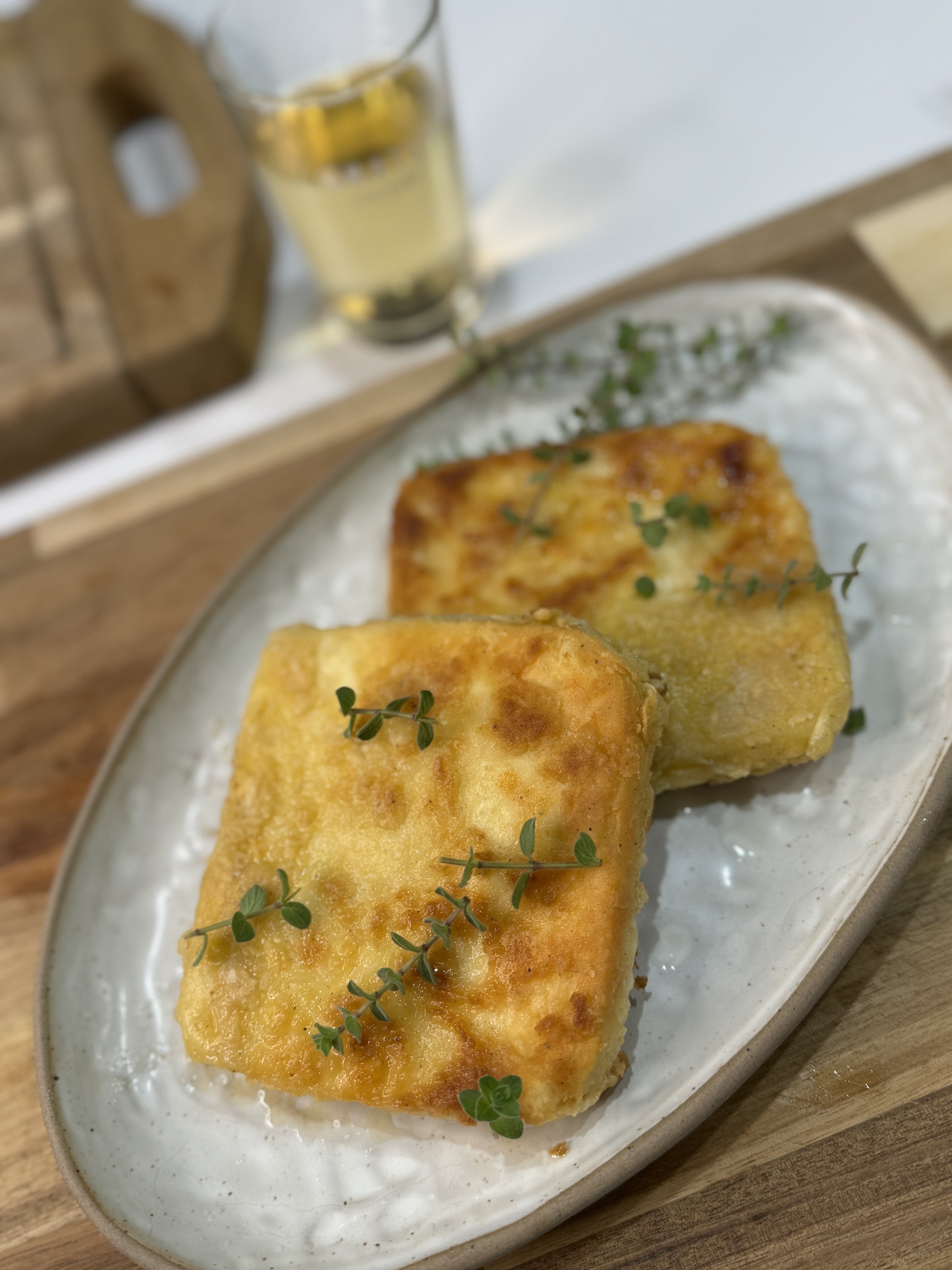 Two golden-brown, breaded square pieces of fried cheese garnished with fresh herbs, served on a white oval plate on a wooden table, with a drink in the background.
